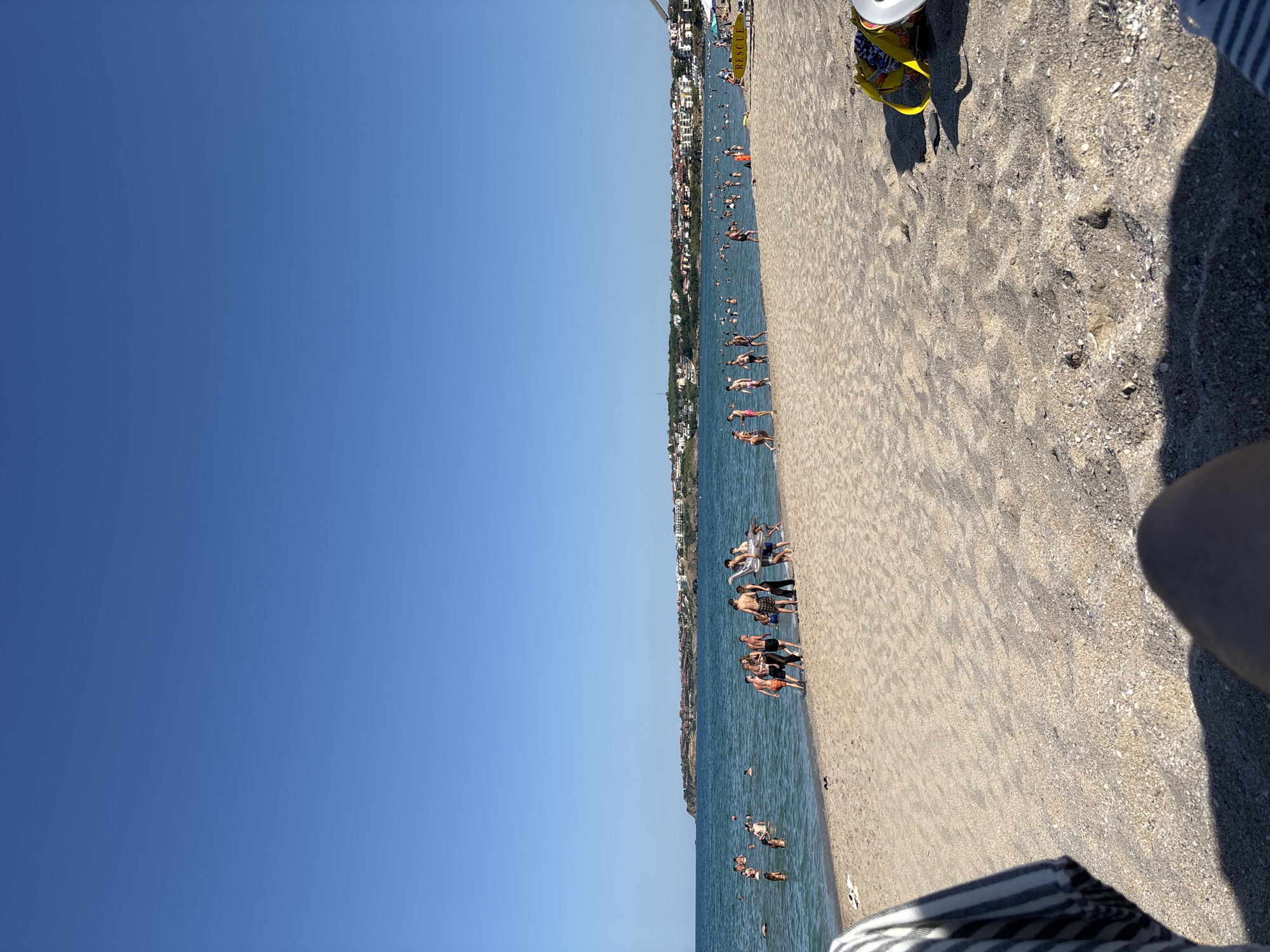 A wide-angle, eye-level shot captures a sunny day at a crowded beach in Sozopol, Bulgaria, with the Black Sea in the background. The foreground is dominated by sand, with a person's leg and a portion of a striped towel visible at the bottom left. In the middle ground, people are scattered across the sand and in the shallow water. Some are gathered in small groups, while others are swimming or wading. A distinctive inflatable dinosaur is visible in the water near the shore. Further out, a line of buildings stretches along the coastline, suggesting a resort town. The sky is a clear, vibrant blue.