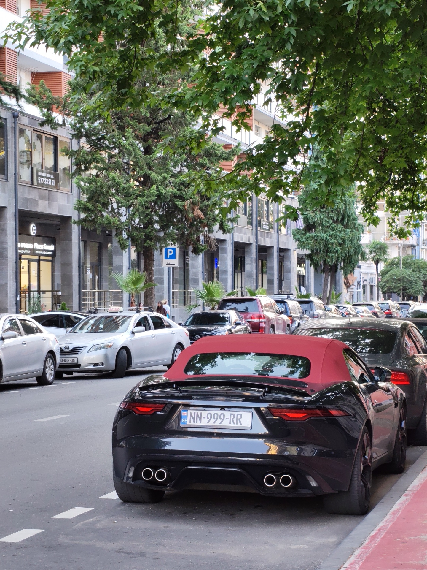 The image shows a street scene in Batumi, Georgia, with several cars parked along the road. In the foreground, a black convertible sports car with a red top is parked. Its license plate reads "NN-999-RR". Further down the street, a silver sedan, possibly a Toyota Camry, is visible with a taxi sign on its roof. Behind it, other vehicles, including a red SUV, are parked. The buildings lining the street appear to be modern with glass storefronts. Trees with green foliage are abundant, casting shadows on the street. A pedestrian walkway with red markings is present along the right side of the road. A blue parking sign is also visible. The overall impression is that of a busy urban environment on a daytime.