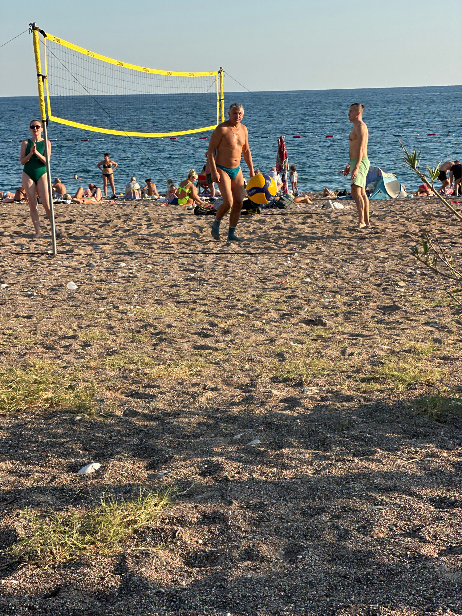 A group of people are on a sandy beach in Boreti, Montenegro, near a volleyball net. In the foreground, a woman in a green swimsuit stands by a volleyball net pole. To her right, a man in blue swim trunks holds a yellow and blue volleyball. Further to the right, another man in green swim trunks stands facing the sea. In the background, other beachgoers are scattered across the sand and in the water, with beach umbrellas and tents visible. The sea is calm and blue, with a clear sky above. The sand is a mix of pebbles and fine grains, with patches of dry grass growing in the foreground.