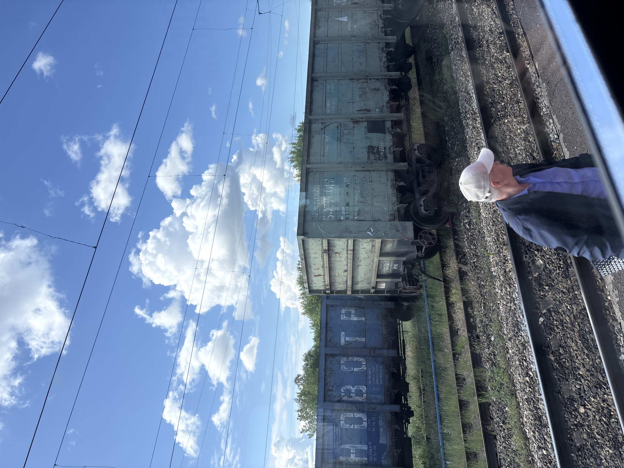 The image depicts a railway scene in Kukmor, Russia, captured from a vantage point, likely a train window. The upper section displays a clear blue sky with scattered white cumulus clouds, crisscrossed by several thin, dark overhead wires. In the midground, two weathered blue-green freight wagons are visible. The wagon on the left bears large Cyrillic lettering "НОВОТР" and discernible smaller text including "ПОСТРОЕН 22" and "15 01 08". The wagon to its right features the number "5477019" and text "ПОСТРОЕН 21" with the date "05.07.2011". A dense line of green trees forms the background above and behind the wagons. The foreground comprises multiple parallel railway tracks resting on a ballast bed, with green grass and weeds growing intermittently between them. A blue pipe or hose runs horizontally along the ground in front of the wagons. In the lower-right corner, an adult male, partially visible due to a window reflection, is depicted wearing a light-colored baseball cap, a light blue shirt, and a dark jacket, looking to the right.