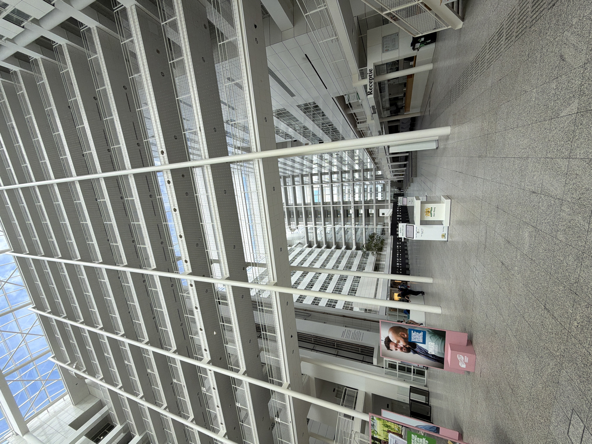 The image shows the interior of a large, modern building with a high atrium. The structure features multiple levels of balconies supported by white columns, with a glass roof allowing natural light to enter. On the ground floor, there is a spacious, tiled area with several displays and a reception desk. Several people are visible walking and standing in the background. Two prominent pink stands display portraits and text. One stand shows two men with text below one of them. Another stand has text and a logo. A sign above a counter reads "Receptie." The overall impression is that of a public or corporate building with an open, airy design. The building is the Ministry of Justice and Security in The Hague, Netherlands.