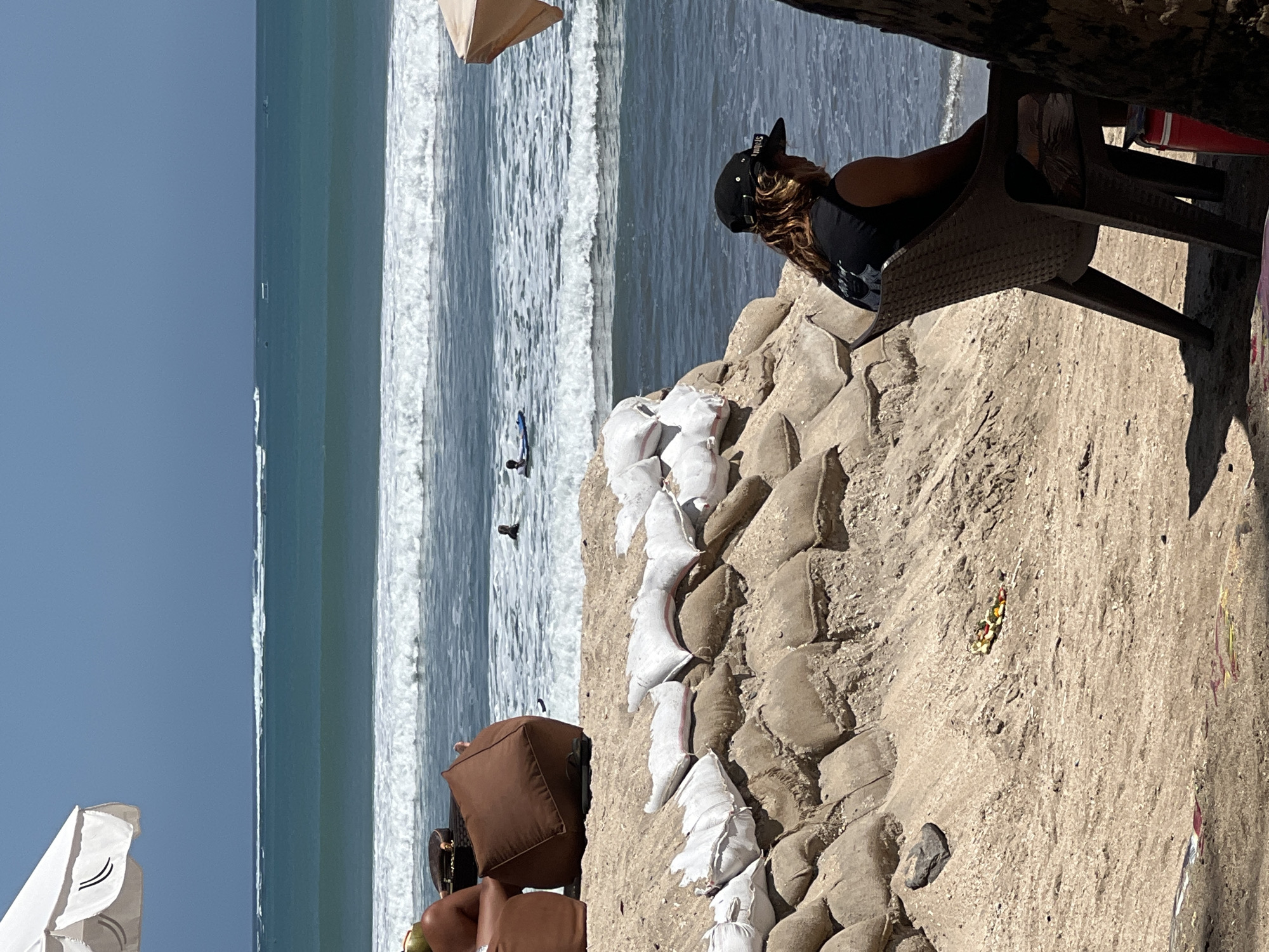 An overhead shot shows a beach with a person sitting on a chair looking at the ocean. The person is wearing a black hat and a black t-shirt. In the background, there are sandbags on the beach and waves crashing in the ocean. The sky is blue and clear. The location is Kuta, Bali, Indonesia.