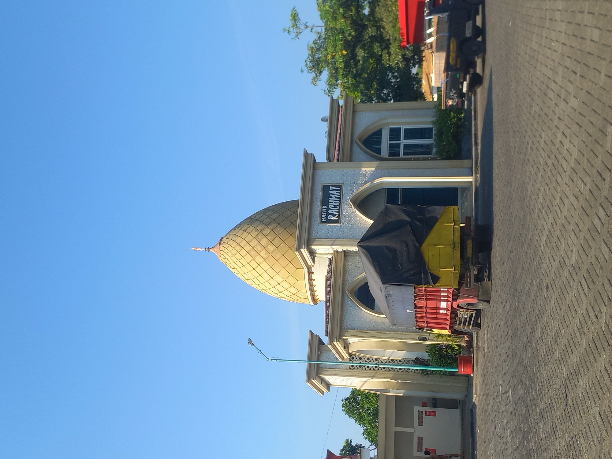 A mosque named Masjid Rachmat, with a distinctive golden dome, stands under a clear blue sky. The mosque features white and cream-colored walls with arched windows and decorative detailing. In front of the mosque, a red and yellow truck with a black tarp covering its cargo is parked on a cobblestone road. To the right of the mosque, a small black pickup truck is visible, also parked. Trees and foliage surround the mosque, and a street lamp stands to its left. A person in casual clothing is partially visible near a building to the far left of the frame. The scene suggests a daytime setting, possibly in a public area with vehicle access.