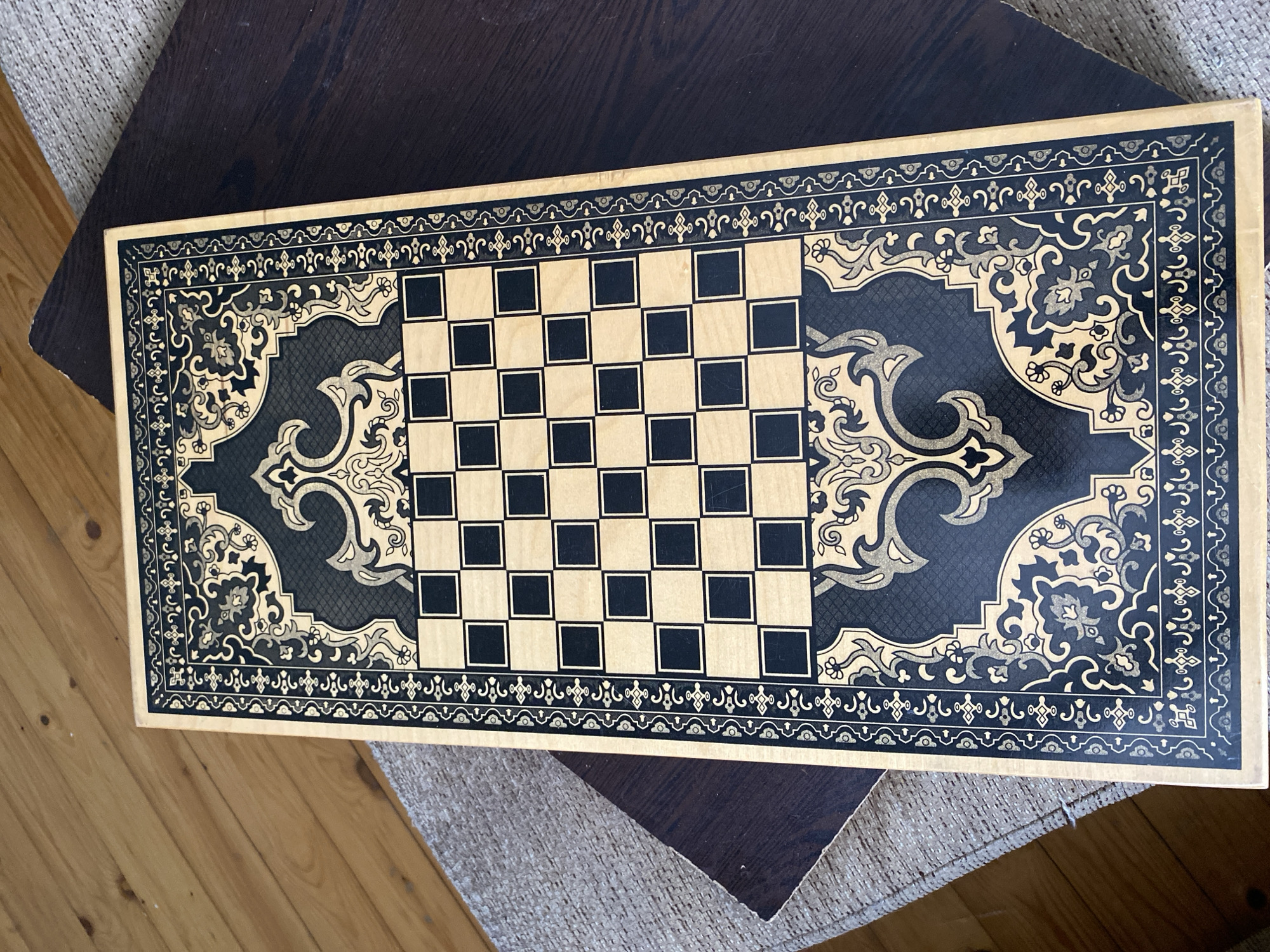 An overhead shot captures a rectangular wooden chessboard with a black and gold ornate border. The chessboard itself is composed of alternating light wood and black squares arranged in an 8x8 grid. The intricate border features geometric patterns and floral motifs in dark blue and gold. The board rests on a dark wooden surface, partially overlapping a textured gray fabric, which in turn lies on a light-colored wooden floor. The scene is well-lit, casting subtle shadows. The image is devoid of any people or animals. The setting is a domestic interior, possibly a living room or study. This image was taken in the city of Bakikhanov, Azerbaijan.
