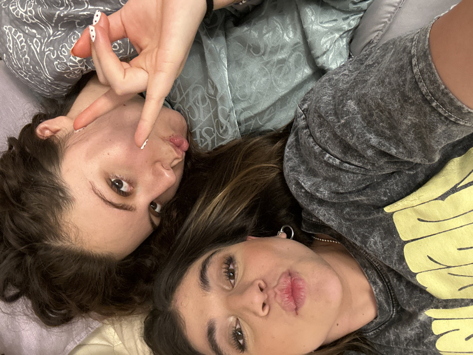 Two young adult females are depicted in a close-up, top-down perspective, appearing to be lying on a soft, light-colored surface. The individual in the lower foreground has long dark hair, her lips pursed, and is wearing a dark grey acid-wash t-shirt with yellow text and a silver hoop earring in her left ear. The individual in the upper background has long dark hair, her head tilted, and her mouth slightly pursed. Her left hand is raised, displaying fingers in a V-sign variant, with nails featuring white tips and small black dots. She is wearing a light blue-green patterned garment. The background appears to be soft bedding or upholstery. The location in the city of Barrio de Nuevo León, Mexico, cannot be visually confirmed from the image content.