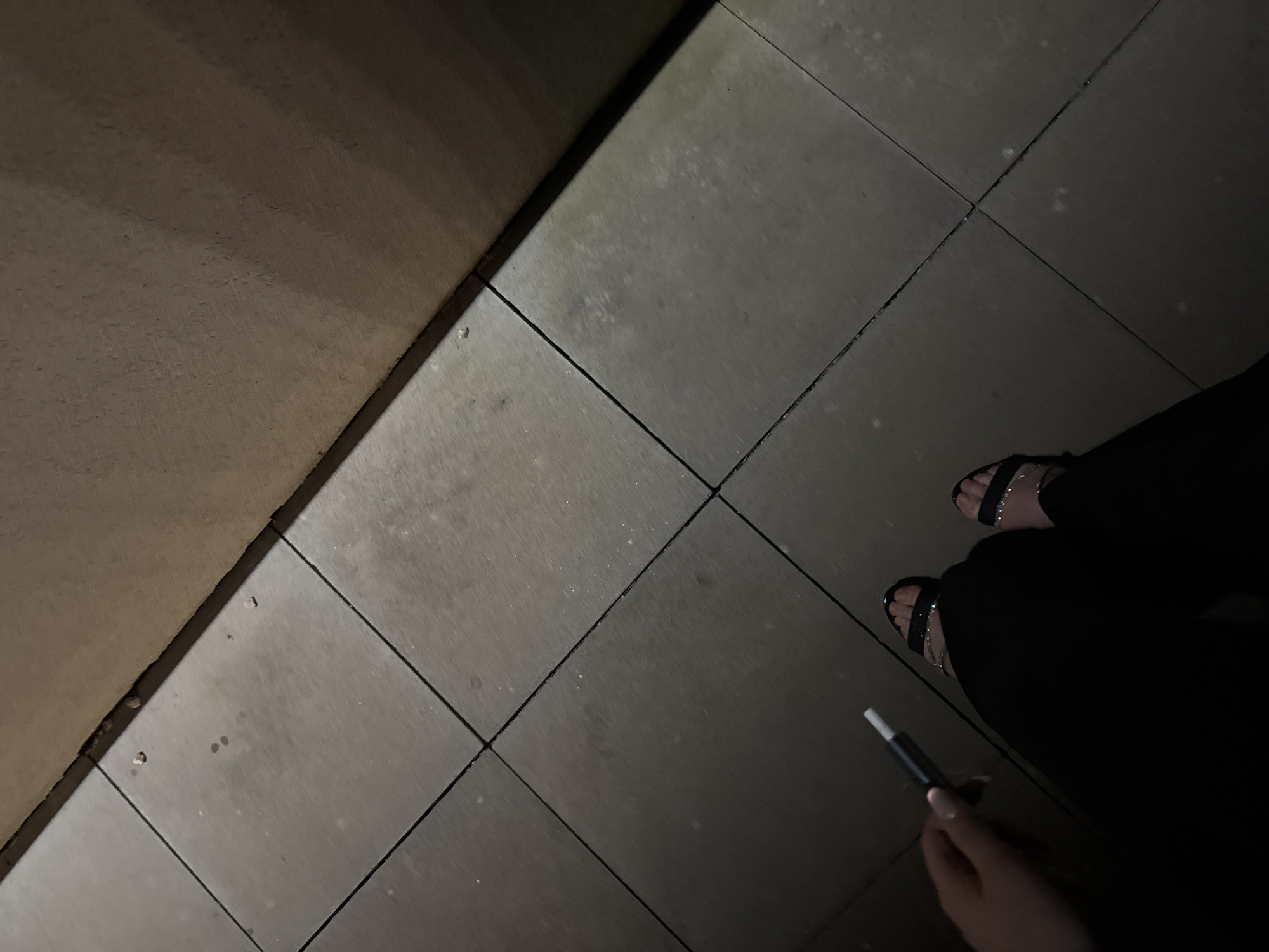 The image depicts a person standing on a paved surface, likely a sidewalk or patio, made of square tiles. The tiles are light grey and show signs of weathering. A low wall or ledge is visible on the left side, its surface a darker shade of grey. The person is wearing black pants and sandals. They are holding a slim, dark-colored object in their hand, possibly a pen or electronic cigarette. The setting is outdoors, and lighting conditions suggest it is nighttime. The location is unknown, and Mogilovo, Bulgaria cannot be determined from the image.