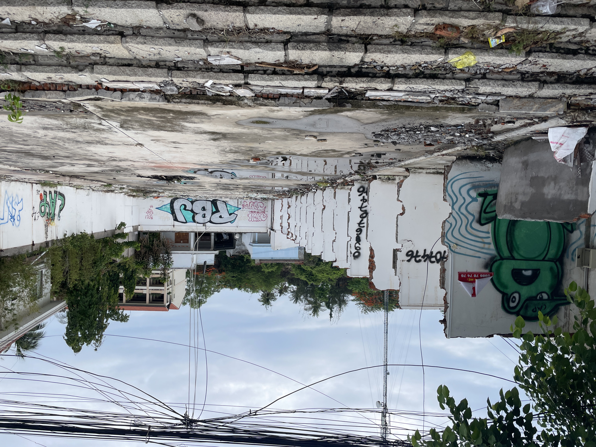 The image depicts an alleyway in Phu Thuy Ward, Vietnam, bordered by damaged buildings with graffiti. The top of the frame shows a crumbling, tiered concrete structure. The walls of the buildings lining the alleyway are white, heavily vandalized with graffiti, and exhibit significant damage, including exposed brick. Vegetation is visible in the middle ground between the buildings, including trees and bushes. Above the buildings, the sky is overcast. Numerous electrical wires crisscross the lower portion of the image, obscuring the view of the sky and buildings behind.