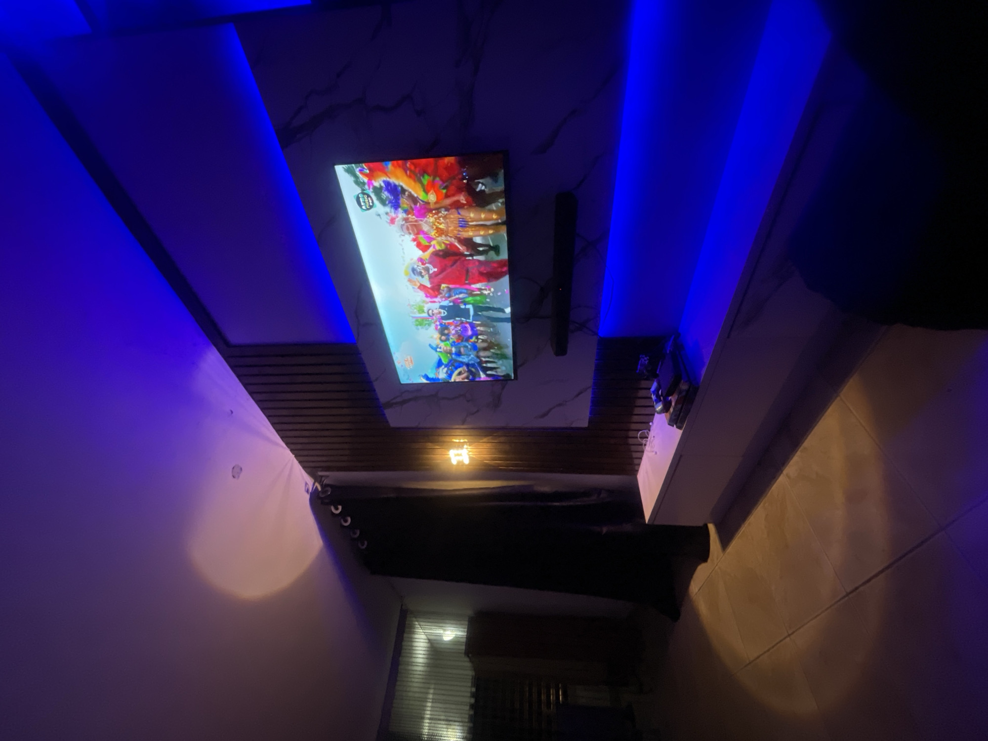 The image shows a room interior with a TV mounted on a wall with a marble-patterned panel. The TV is displaying a colorful image. Above the TV, there is a ceiling design with blue LED lighting. A curtain hangs to the side of the wall. Below the TV, a light fixture shines. There's a shelf or surface with objects on it to the right of the TV. The room has a tiled floor. The lighting appears to be a combination of natural light filtering through a window in the distance, and artificial, blue-toned light. The location is unknown.
