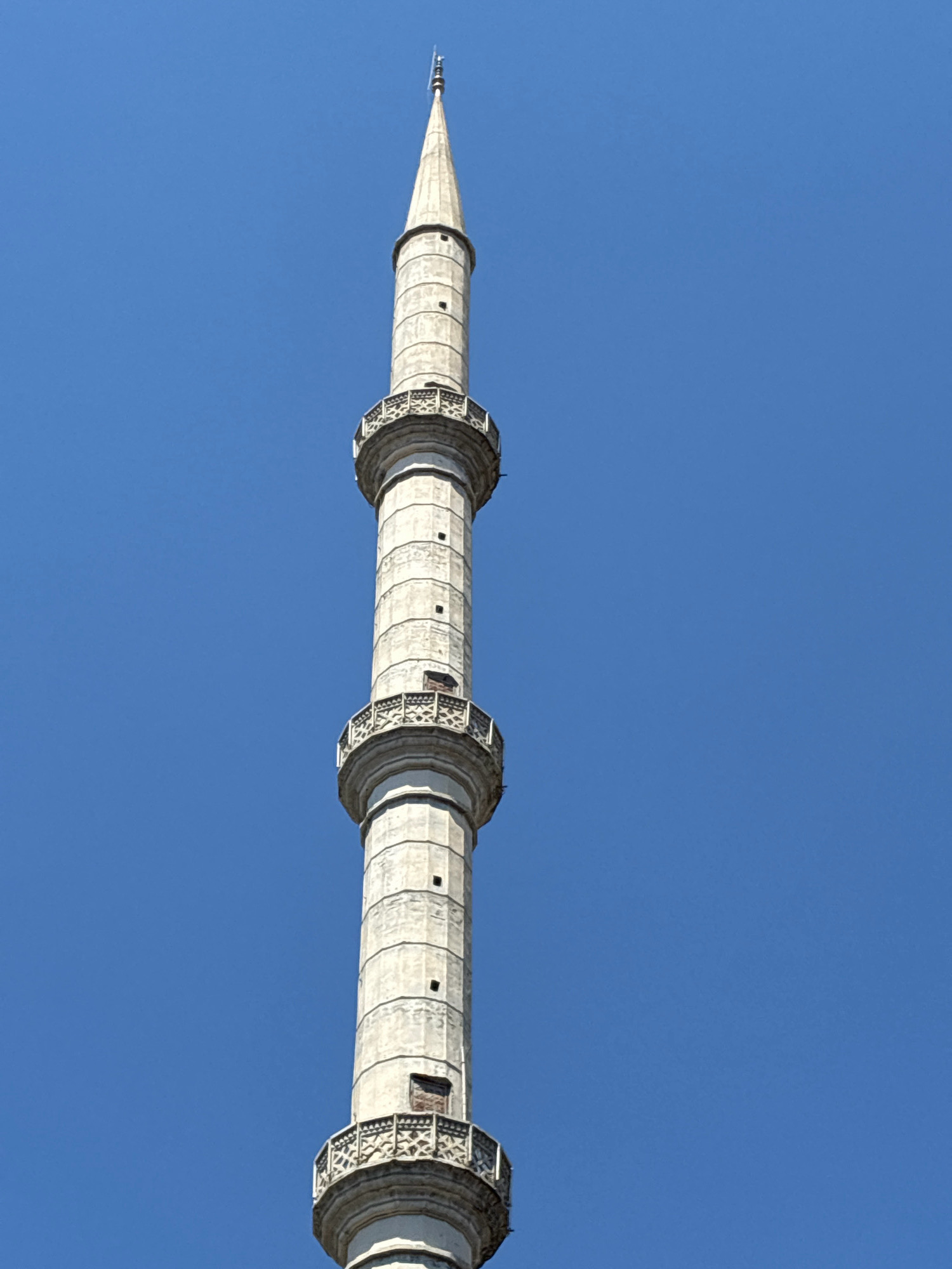 The image shows a close-up, low-angle view of the minaret of the Red Mosque (Kızıl Minare) in Alanya, Turkey, set against a clear, vibrant blue sky. The minaret is tall and slender, constructed from light-colored stone. It features a series of stacked cylindrical sections, separated by decorative bands. Three distinct balconies, or *şerefes*, are visible along the height of the minaret. These balconies have intricate, patterned railings and project outward from the main structure. Small, rectangular openings, likely windows or decorative elements, are present at various levels of the minaret. The topmost section tapers to a pointed spire, capped with a finial. The overall condition of the minaret appears weathered but intact, showcasing architectural details of historical significance. The environment is solely the sky, offering no context of the surrounding city or ground level. There are no people or other objects visible in the frame.