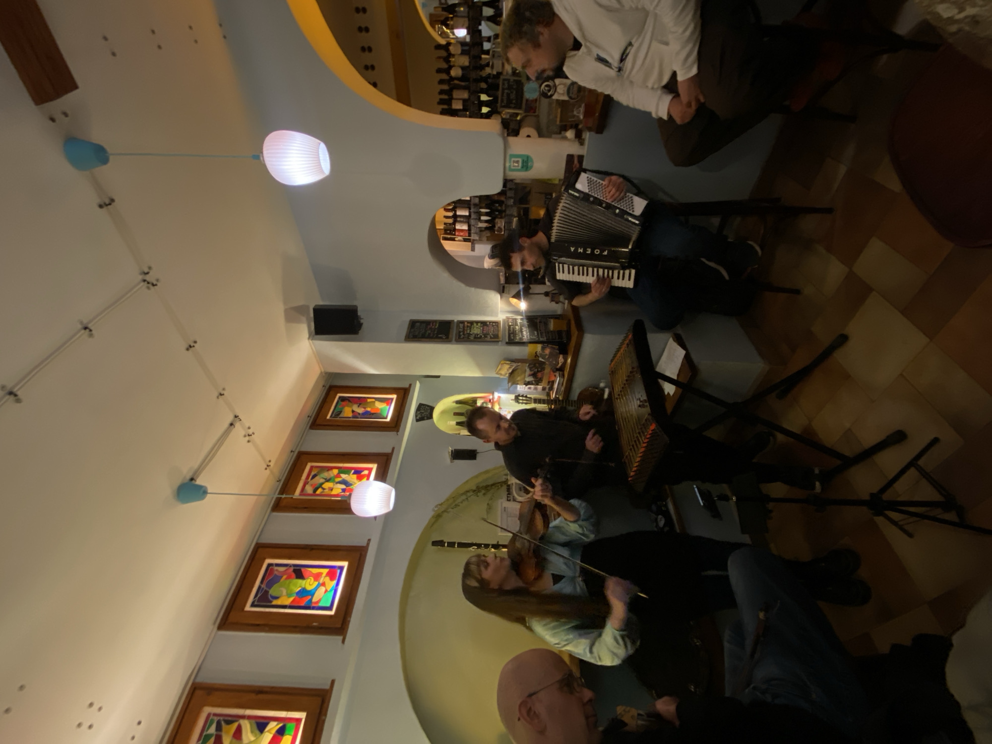 A group of musicians performs in a venue that appears to be a bar or restaurant in Sofia, Bulgaria. In the background, shelves are stocked with bottles, suggesting the location is a bar. To the left, four framed paintings are hung on the wall. A woman is playing the violin, another man plays a dulcimer, and another man is playing the accordion. In the foreground is the back of a man's head. Overhead is a string of pendant lights. The musicians are seated, and additional people are sitting at the bar and at tables. The floor is tiled.