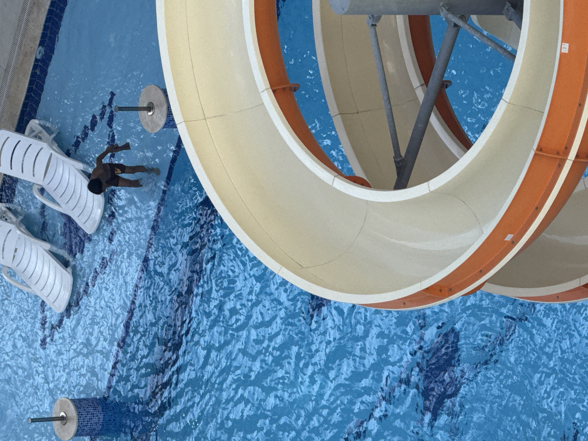 The media file presents an overhead, high-angle view of a vibrant swimming pool scene, likely at a resort in Alanya, Turkey. The central and right portions of the frame are dominated by a large, curving water slide, which features a cream-colored inner surface and a contrasting orange exterior. Grey metal support beams for the slide are also visible.
Below the water slide and extending across the frame, the swimming pool's blue water shimmers with ripples and reflections, revealing darker, patterned tiles on the pool floor. In the left-middle section of the pool, a person, appearing to be a child or young adult, is partially submerged in the water, floating or gently swimming. They are wearing dark shorts. Nearby, two white, slatted plastic lounge chairs rest at the edge of the pool.
Two cylindrical pool fixtures, one partially covered in blue mosaic tiles, are visible in the water, each secured by a metal rod protruding upwards. The scene takes place during a bright, sunny daytime, indicated by the clear reflections on the water and overall illumination, suggesting a recreational and leisurely atmosphere. No legible text is visible on any of the objects within the image, though small white rectangular labels are present on the orange sections of the water slide.