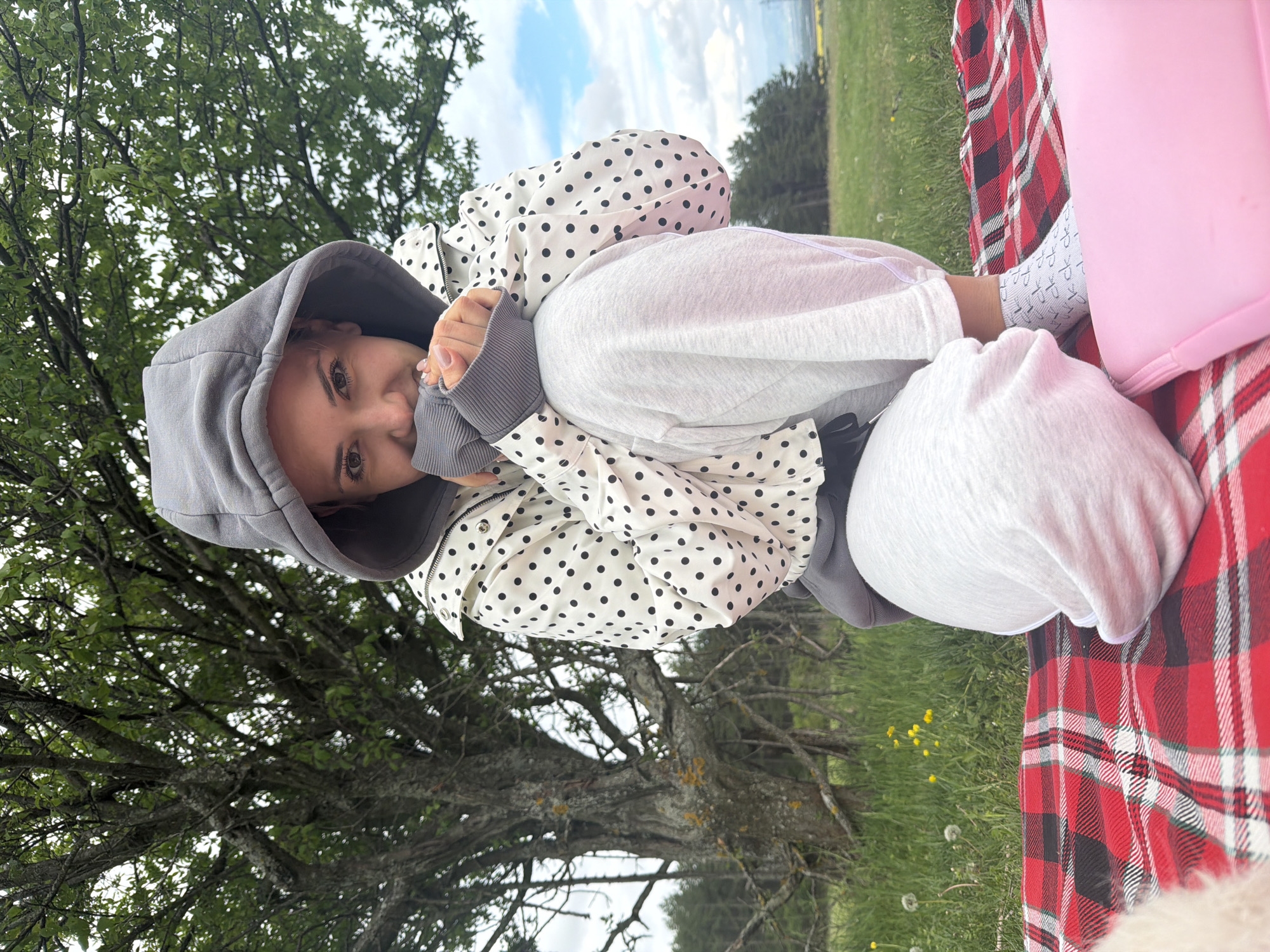 The image shows a person outdoors, likely in a park or grassy area, possibly in Sofia, Bulgaria, given the prompt. The person is sitting on a red plaid blanket. They are wearing a gray hooded sweatshirt and a white jacket with black polka dots. They are looking at the camera and have their hands near their face. A large tree is visible behind the person. The sky is visible, with some clouds. The overall scene suggests a casual or relaxed setting, potentially a picnic or outdoor gathering. A pink object is also on the blanket.