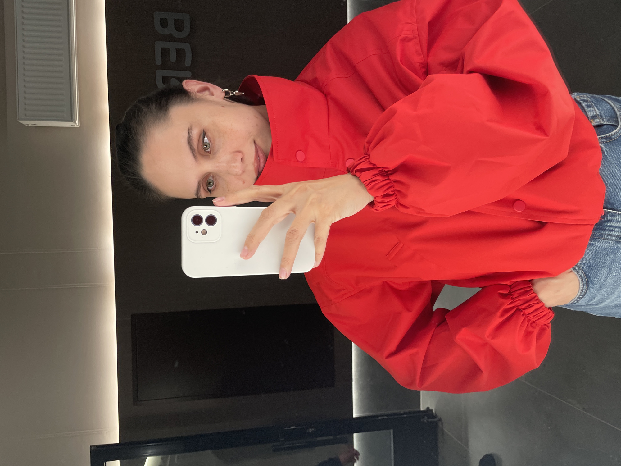 A woman is taking a selfie indoors. She is wearing a red jacket and jeans, and holding a white smartphone. The background includes a dark wall with the word "BEL" partially visible, a vertical light fixture, and a dark television screen. The location is Izhevsk, Russia.