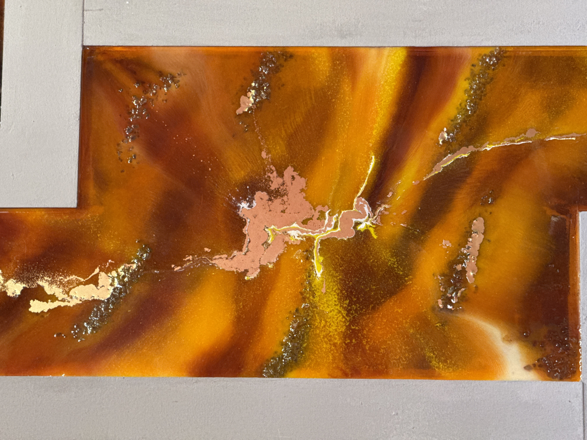 The image shows a rectangular artwork with an orange, brown, and yellow abstract design, framed by a grey border. The central portion features a burst pattern with gold and copper-colored accents scattered throughout. Small clusters of dark, possibly metallic particles are embedded within the surface. There is no indication of the location within Taganrog City District, Russia.