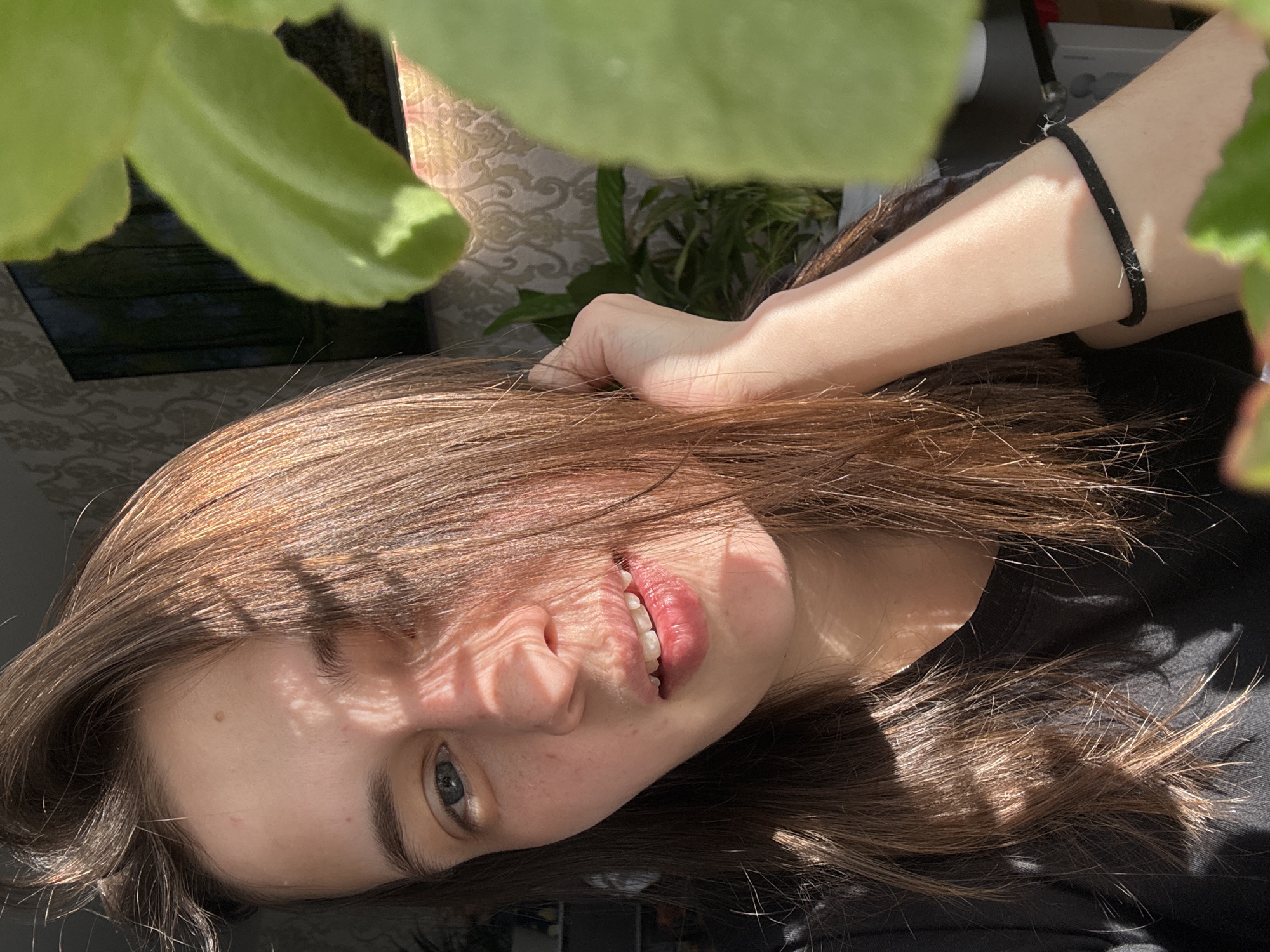 A close-up, horizontally oriented image features a young person, likely female, lying down in bright sunlight. Her head is tilted to the right, and she looks upwards with a soft, gentle smile, her lips slightly parted to reveal white teeth. Her eyes are light blue or grey.
She has long, straight brown hair, which appears to be highlighted with lighter tones by the sun. Strong, directional sunlight casts distinct shadow patterns, possibly from blinds or plant leaves, across her face, particularly on her forehead, nose, and cheek, creating stripes of light and shade. Small freckles are visible across her nose and cheeks, and a small mole is present above her left eyebrow.
Her right hand is visible, resting in her hair near her temple. On her left wrist, a simple black hair tie, appearing slightly worn, is visible. She wears a dark, possibly black, casual top.
The setting appears to be indoors, with a patterned wallpaper or fabric visible in the background. A dark object, possibly a plant pot, is present behind green foliage. Several large, bright green plant leaves are prominently in the foreground, partially obscuring the top and bottom edges of the image, and likely contributing to the shadows on her face. The bright sunlight suggests it is daytime. The overall mood is relaxed and natural. There is no visible text in the image.