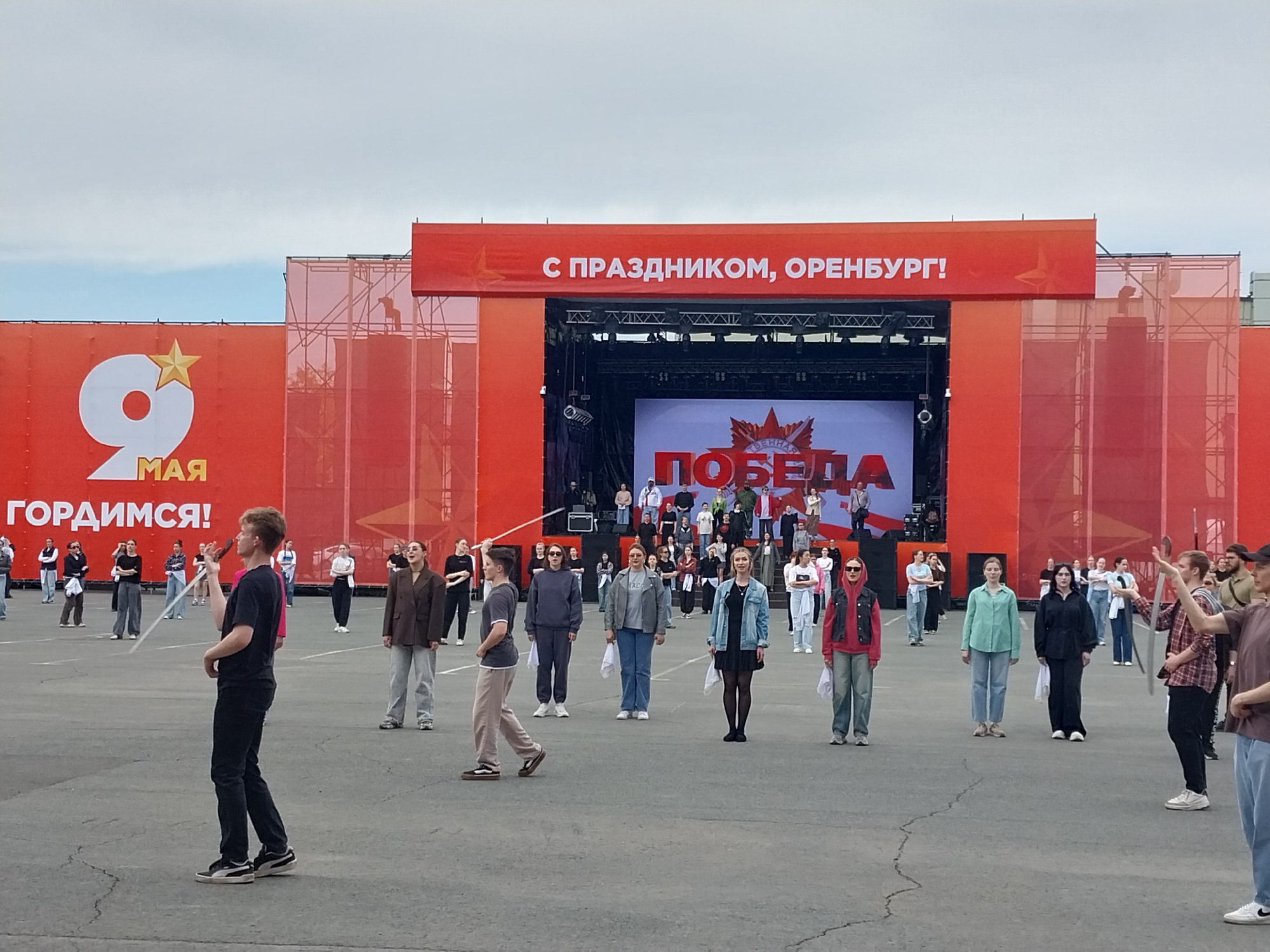 The image shows a public gathering in what appears to be Orenburg, Russia, based on the prominent banner overhead that reads "С ПРАЗДНИКОМ, ОРЕНБУРГ!" (Happy Holiday, Orenburg!). Several individuals are standing in rows on a gray concrete surface, possibly a town square or designated outdoor area. In the background, there is a large stage or platform with red panels on either side and a backdrop displaying the Russian word "ПОБЕДА" (Victory), which suggests the event is related to Victory Day celebrations on May 9th, which are also seen on a sign in the image "9 мая гордимся!". Most of the individuals are facing the stage and holding small white flags or cloths. The people's attire ranges from casual to slightly more formal, with jackets, jeans, and dresses being visible. A few individuals are holding microphones, possibly preparing to speak or perform. The environment suggests a moderately overcast day, with the sky appearing gray.