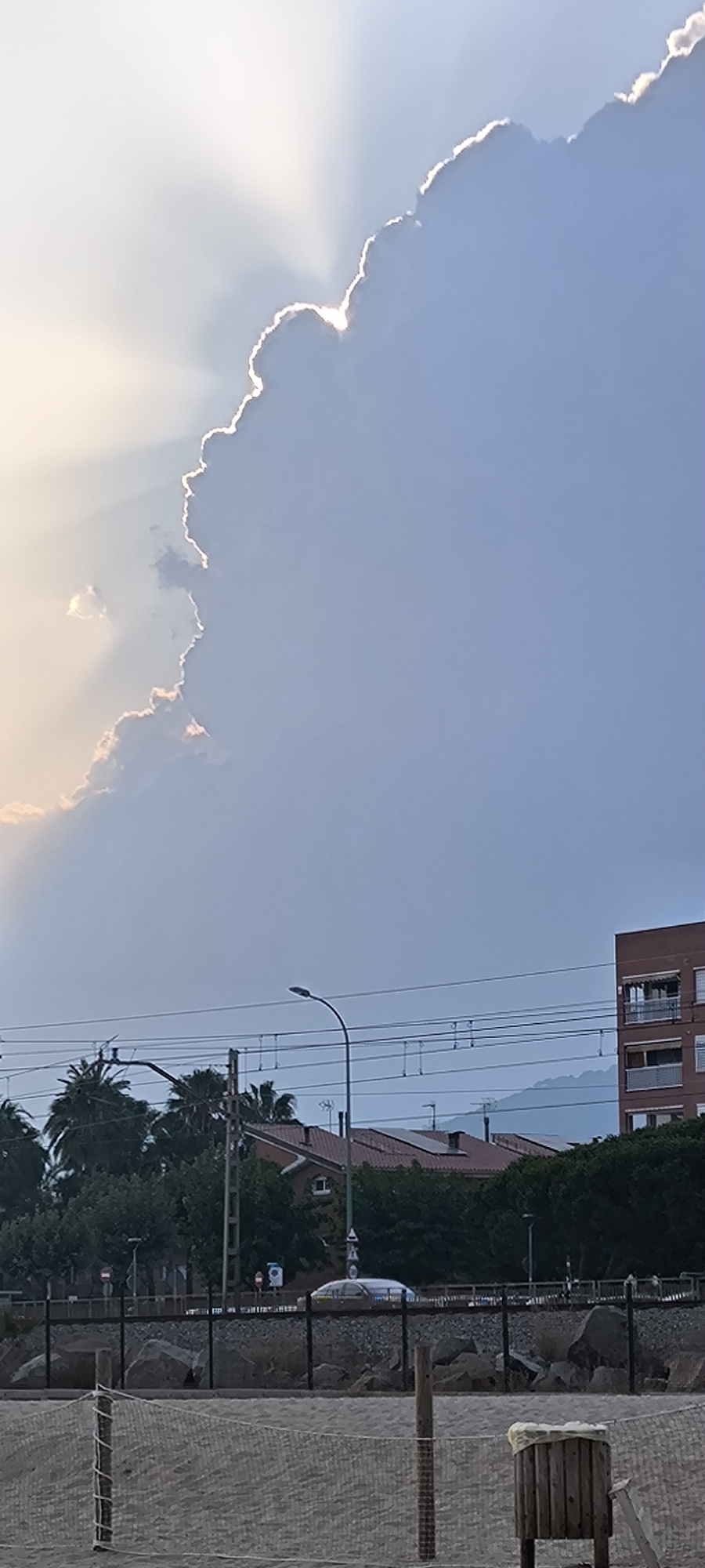 The image depicts a beach scene in Premià de Mar, Spain, at what appears to be either sunrise or sunset, given the dramatic lighting. A large, illuminated cloud dominates the upper portion of the frame, with rays of light piercing through its edges, creating a radiant effect against a pale blue sky.
In the middle ground, a street light with a curved arm is visible, along with overhead power lines and their supporting structures. Behind these, a cluster of buildings can be seen. To the left, palm trees are interspersed with lush green foliage. Further to the right, a reddish-brown multi-story building with balconies stands out, and a dark green line of trees forms a dense backdrop. Rooftops of several buildings are visible, some appearing to have solar panels.
The foreground consists of a sandy beach. A net fence with wooden posts stretches across the lower part of the image, demarcating an area of the beach. In the far right of the foreground, a wooden trash receptacle is positioned. A section of a wall made of large rocks is visible behind the fence, separating the beach area from what appears to be a pathway or road where a light-colored vehicle is present. The overall scene captures a coastal urban environment with a prominent natural sky phenomenon.