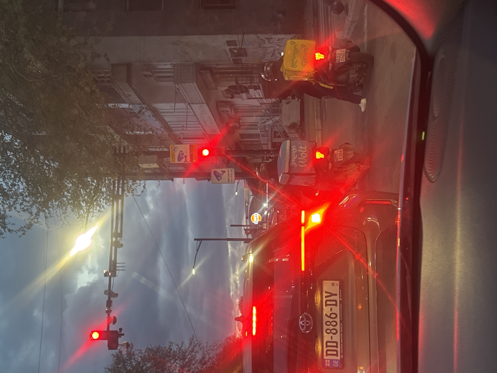 The image shows a street scene in Tbilisi, Georgia. The view is from inside a car, looking out the front windshield at an intersection at a red light. A Toyota car with license plate "DD-886-DY" is in front of the camera car. Two delivery motorcycles with food delivery bags are also in the scene. One has a "Glovo" bag, with license plate "EJJ 0603". The other has a "Wolt" bag, with license plate "ETT 8266". A Gulf gas station sign is visible in the background. Buildings line the street.