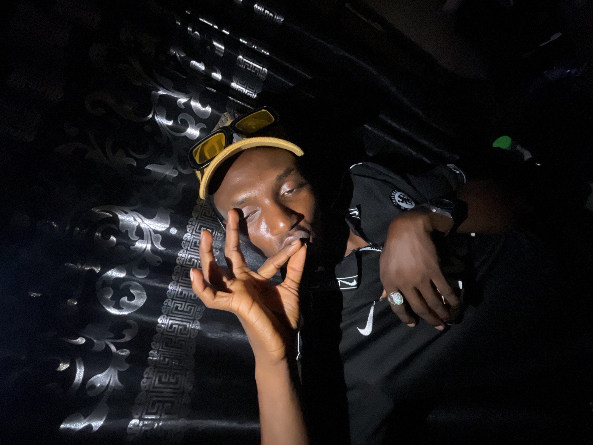 A man is lying down on what appears to be a black patterned textile. He is wearing a black t-shirt with a white Nike logo and what appears to be a Chelsea FC emblem. He has on a yellow baseball cap with yellow sunglasses perched on top. His hand is raised to his mouth, and he is wearing a ring on his right hand. The background is dark, and the setting could be indoors. The location in the city of Lokoja, Nigeria is not discernible.