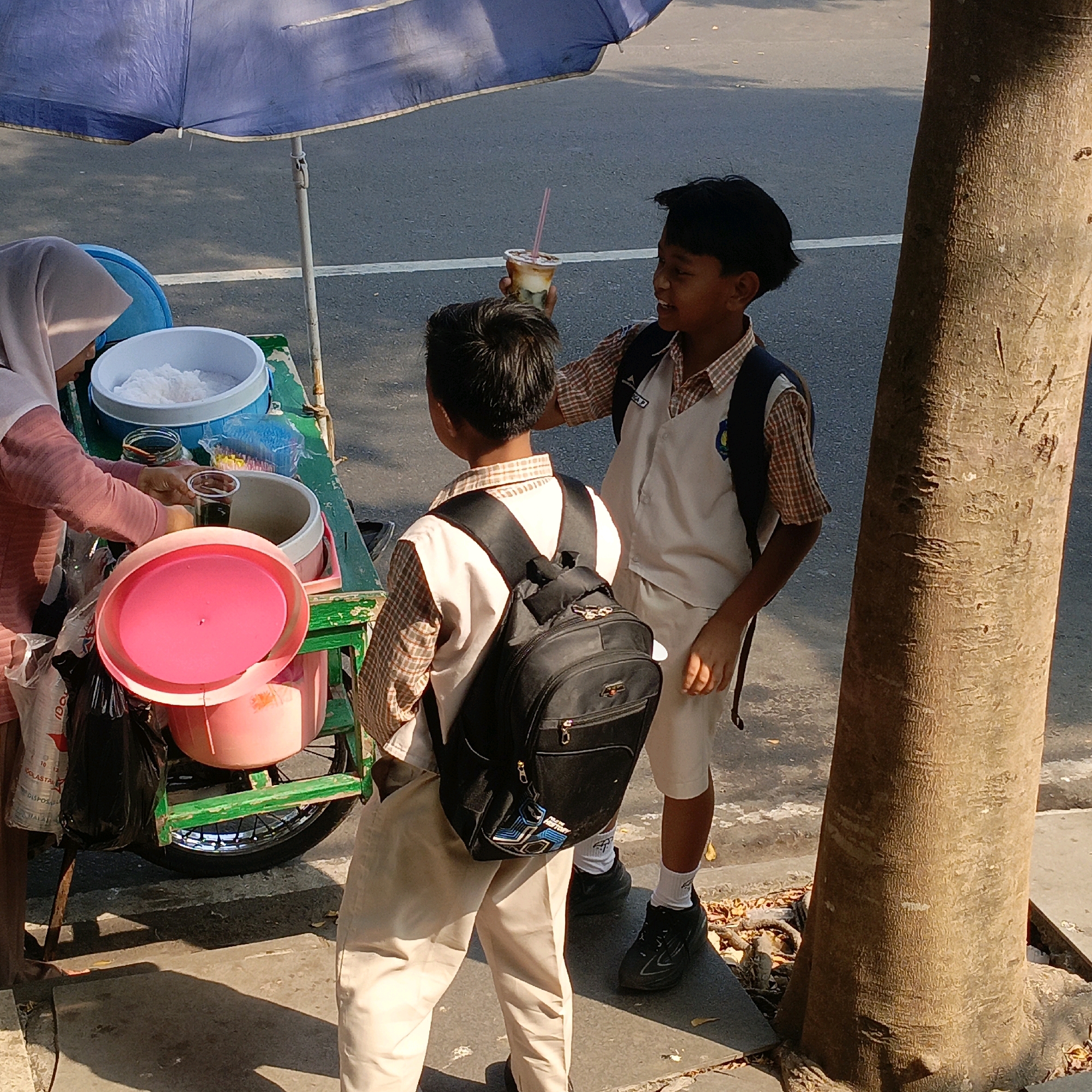 In Salatiga, Indonesia, a street vendor selling drinks is partially visible from the chest down, wearing a pink hijab and a pink top. She is standing behind a green cart filled with containers of ice and drinks. Two young boys, appearing to be students in uniform with beige pants and white shirts with plaid collars, are interacting with the vendor. One boy, seen from the back, wears a black backpack. The other boy, facing the vendor, holds a cup with a straw and is reaching out his hand. They are positioned on a paved surface next to a large tree. The scene is outdoors, under a blue umbrella providing shade.