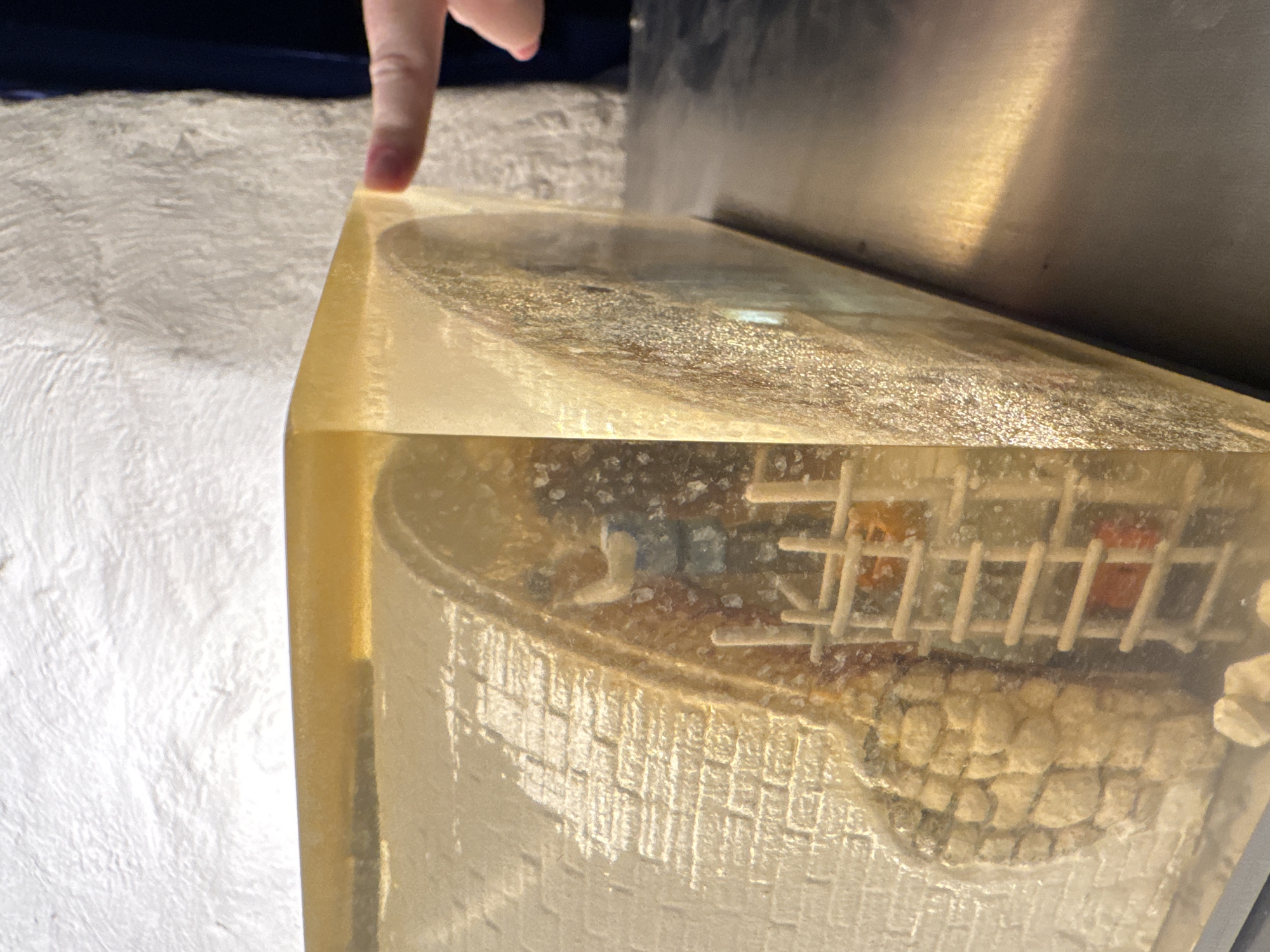 A person's index finger rests on the corner of a translucent, rectangular block of resin. Encased within the resin is a miniaturized model of a building. The model shows a domed structure with intricate brickwork details on its lower section and what appears to be a grid-like upper section. The resin block rests on a textured, off-white surface. A dark blue shape is visible behind the finger. The top of the resin block has a grainy texture. The presence of the domed structure and the brickwork suggests the Kul Sharif Mosque, a landmark in Kazan, Russia, is depicted within the resin block.
