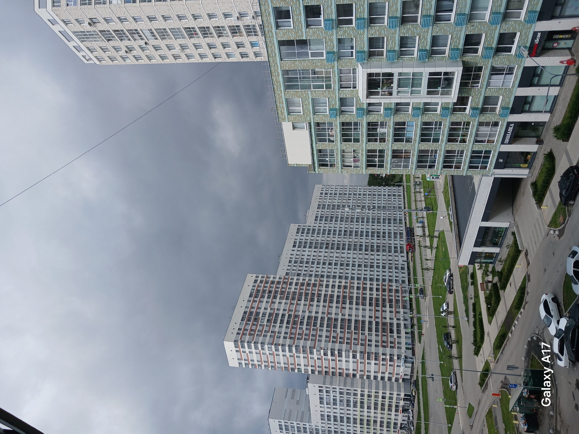A high-angle, medium shot captures a residential area in Yekaterinburg, Russia, under a cloudy sky. Several multi-story apartment buildings dominate the scene. In the foreground on the right is a building with a light-green tiled facade and numerous windows. To the left, a taller white and orange striped apartment building stands prominently. Further in the distance to the left, another white apartment building with a pattern of grey rectangles is visible. The ground level of the apartment buildings in the center and right features storefronts. Paved roads with parked cars and green spaces with trees and bushes are present. One building in the center has a sign reading "ПИВНАВКА," indicating a store. The sky is overcast with dark grey clouds. A thin wire is visible diagonally across the upper portion of the image. The bottom left corner of the image displays "Galaxy A17."