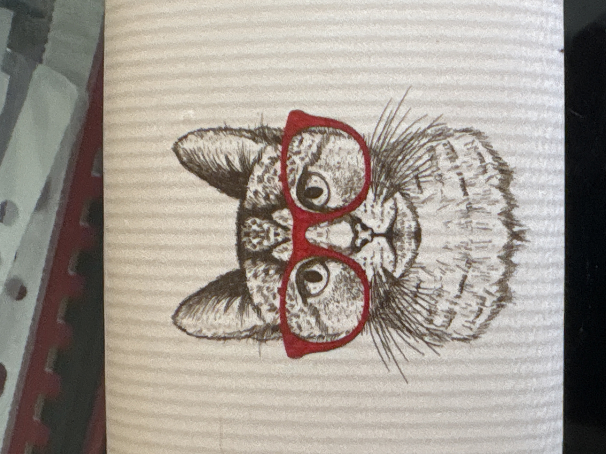 The image shows a detailed sketch of a cat wearing red glasses. The cat is facing forward. The sketch is on a light background with horizontal lines. Other elements include a red shelving unit and a black object in the foreground. The location is not discernible from the image.