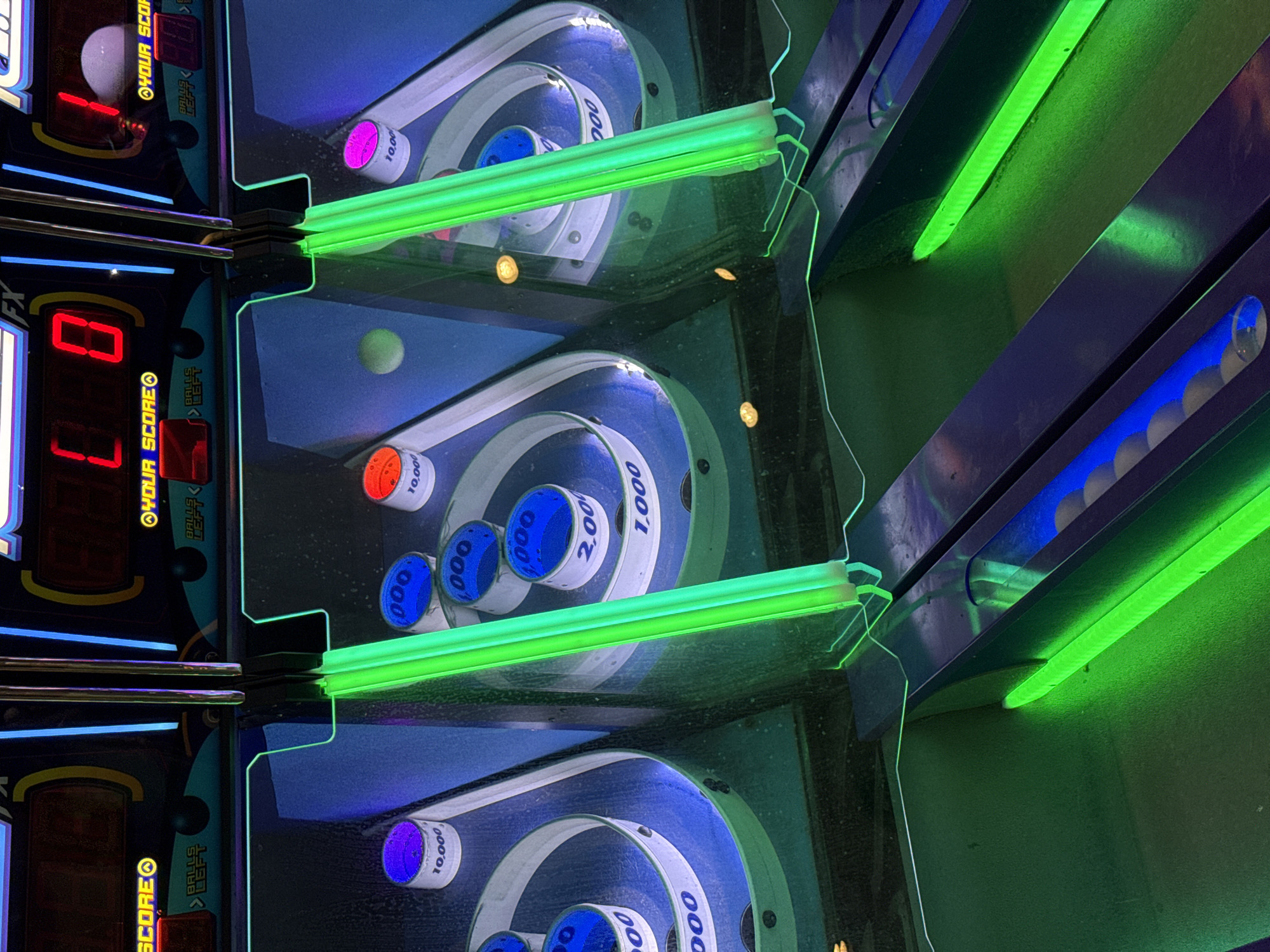 The image captures a close-up, angled view of a skee-ball game, an arcade activity where players roll balls up a lane to score points by landing them in numbered holes. The image is dominated by the blue and white scoring rings, each labeled with point values such as 1,000, 2,000, and 10,000. A green fluorescent light bar illuminates the playing surface, adding to the arcade atmosphere. The digital scoreboards on the left show the current score as 0. The precise location within the city of Columbus, United States of America, is not identifiable from the image.