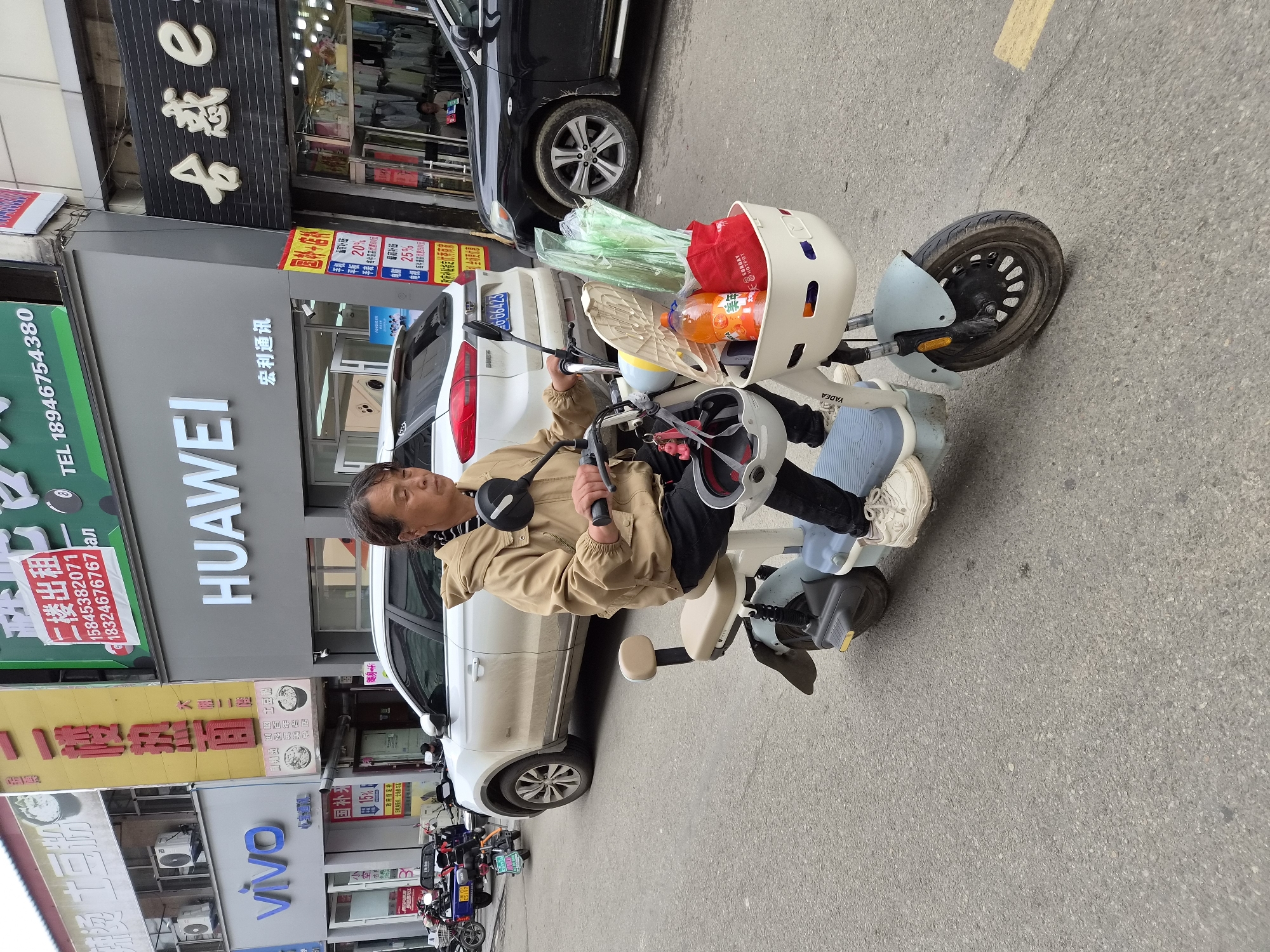 A woman is riding a light blue electric scooter down a street in Mishan, China. She is wearing a tan hooded jacket, black pants, and white sneakers. She has a white basket attached to the front of the scooter, which contains a bottle of orange drink, a red bag, and some green plastic bags. A white car is parked behind her, and a black car is to her right. Various businesses with Chinese signage are visible on the buildings lining the street.