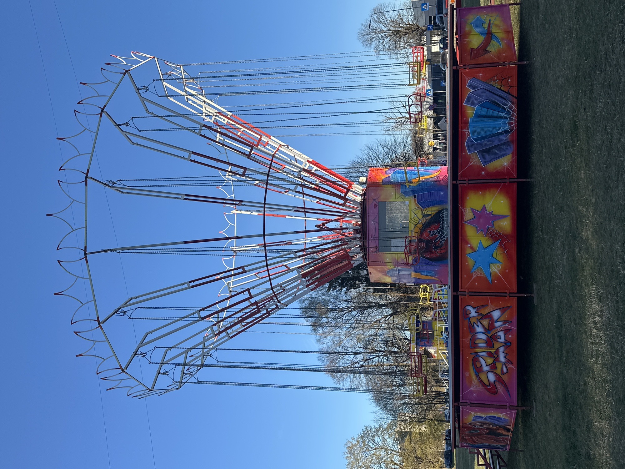 This media file presents a vibrant swing ride, commonly known as a chain carousel, situated outdoors in what appears to be a fairground or park setting in Riga, Latvia. The ride dominates the left side of the frame, showcasing its intricate structure against a clear, bright blue sky.
The main subject is a large, circular swing ride with numerous chains hanging down, awaiting seats for riders. The ride's framework consists of white and red metal beams, forming a complex canopy from which the chains descend. Decorative arched elements with red tips adorn the outer edge of this structure.
The base of the ride, visible on the right, is a colorful structure adorned with graffiti-style artwork. Adjacent to the ride is a barrier or fence composed of panels, also brightly painted with similar artistic themes. One panel clearly displays the text "SPIDER MAN" in stylized red, orange, and white letters, accompanied by spiderweb designs, suggesting a superhero theme. Other panels feature a blue star, abstract patterns, and additional spiderweb motifs.
The ride appears to be stationary, with no people visible on or around it, suggesting it might not be in operation at the moment, or perhaps it's being set up or dismantled. The image captures bright daylight under a cloudless blue sky. In the background, bare deciduous trees are visible, indicating either early spring or late autumn. The ground surrounding the ride is grassy. A pedestrian crossing sign can be seen faintly in the distant background, hinting at proximity to a road or public access area.