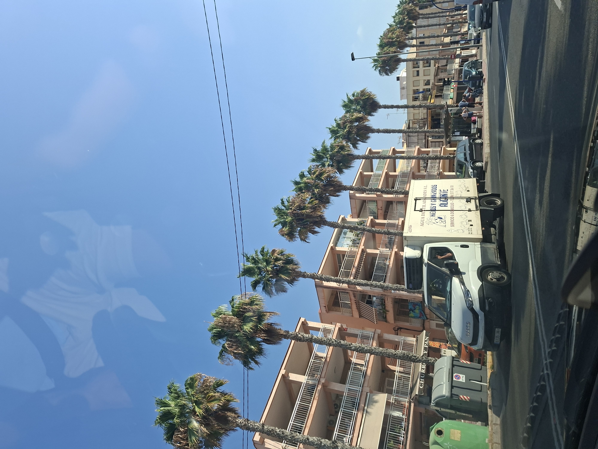The image depicts a street scene in Torrevieja, Spain, on a clear, sunny day. In the foreground, a white Iveco delivery truck is traveling on a multi-lane road. The truck has a refrigeration unit on its roof and signage on its side that reads "AGUA DESTILADA Y HIELOS GANIZADOS ALICANTE SERVICIO A DOMICILIO". To the left of the truck are two large, commercial-sized trash bins, one green and one gray. Behind the truck and trash bins, a row of apartment buildings with pale peach-colored facades lines the street. These buildings feature multiple balconies with white railings. Several tall palm trees with green fronds are interspersed along the sidewalk in front of the buildings, casting shadows on the road. Further down the street, more apartment buildings and shops are visible, and a street lamp stands on the right side. The sky is a clear, bright blue, with some faint reflections visible on what appears to be a car windshield, suggesting the photo was taken from inside a vehicle. A few people are visible on the sidewalk near a bus stop or shelter.