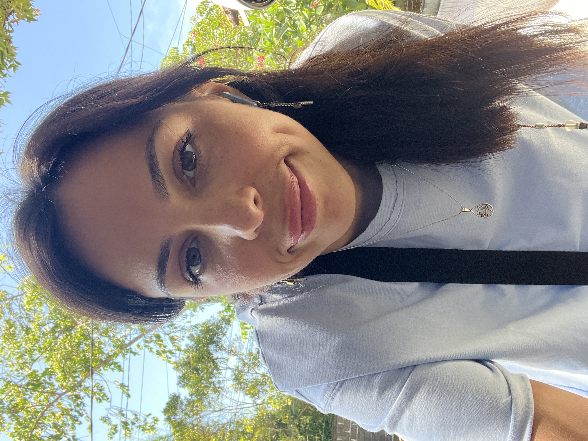 A woman poses outdoors in Kuta Selatan, Indonesia, on a sunny day. The woman is wearing a light blue long-sleeved shirt and a silver necklace with a pendant. She has dark brown hair and brown eyes. A black strap is visible across her chest. In the background, trees and a clear blue sky are seen. A silver key-shaped object and beaded hair accessories are visible in her hair. The scene is a casual, candid shot.