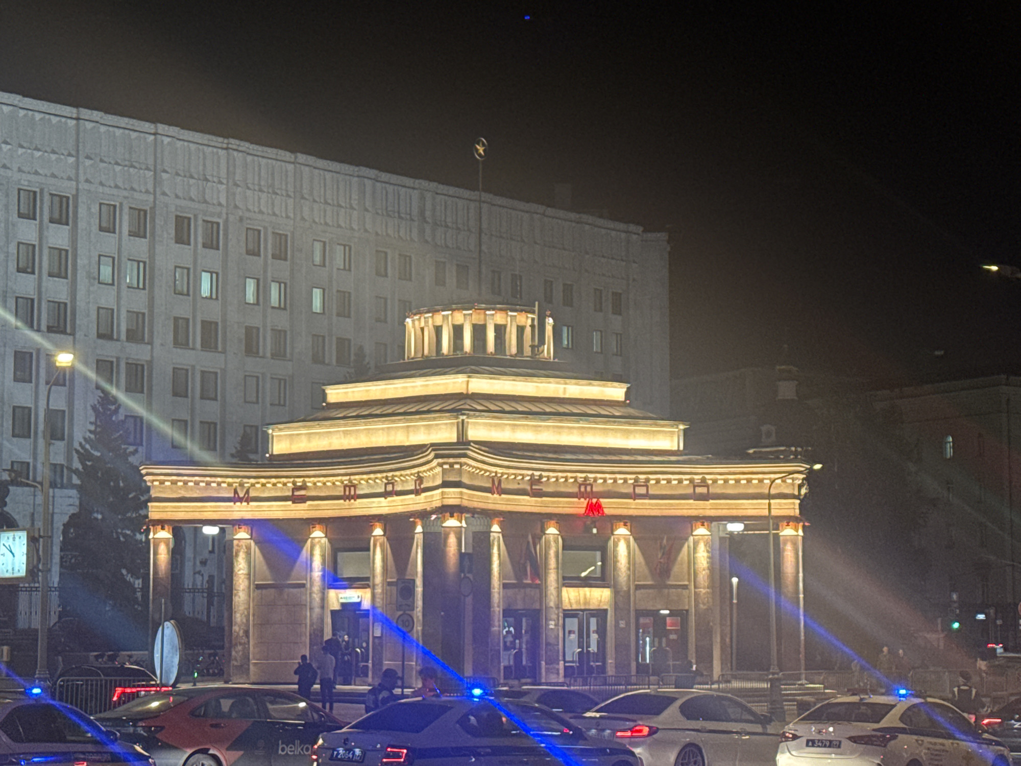 The image shows a night scene in Moscow, Russia, featuring the iconic Metro station entrance of Mayakovskaya. The station's Art Deco architecture is illuminated with warm, golden lights, highlighting its circular dome and colonnade. Red Cyrillic lettering spelling "МЕТРО" is prominently displayed above the entrance. In front of the station, several vehicles are visible, including what appear to be police cars with flashing blue lights, suggesting an active or recent event. People are seen near the entrance and on the sidewalk. Behind the Metro station, a large, imposing building with numerous windows is visible, its facade illuminated by streetlights. The overall atmosphere is one of a bustling urban environment at night, with a mix of architectural grandeur and contemporary activity.