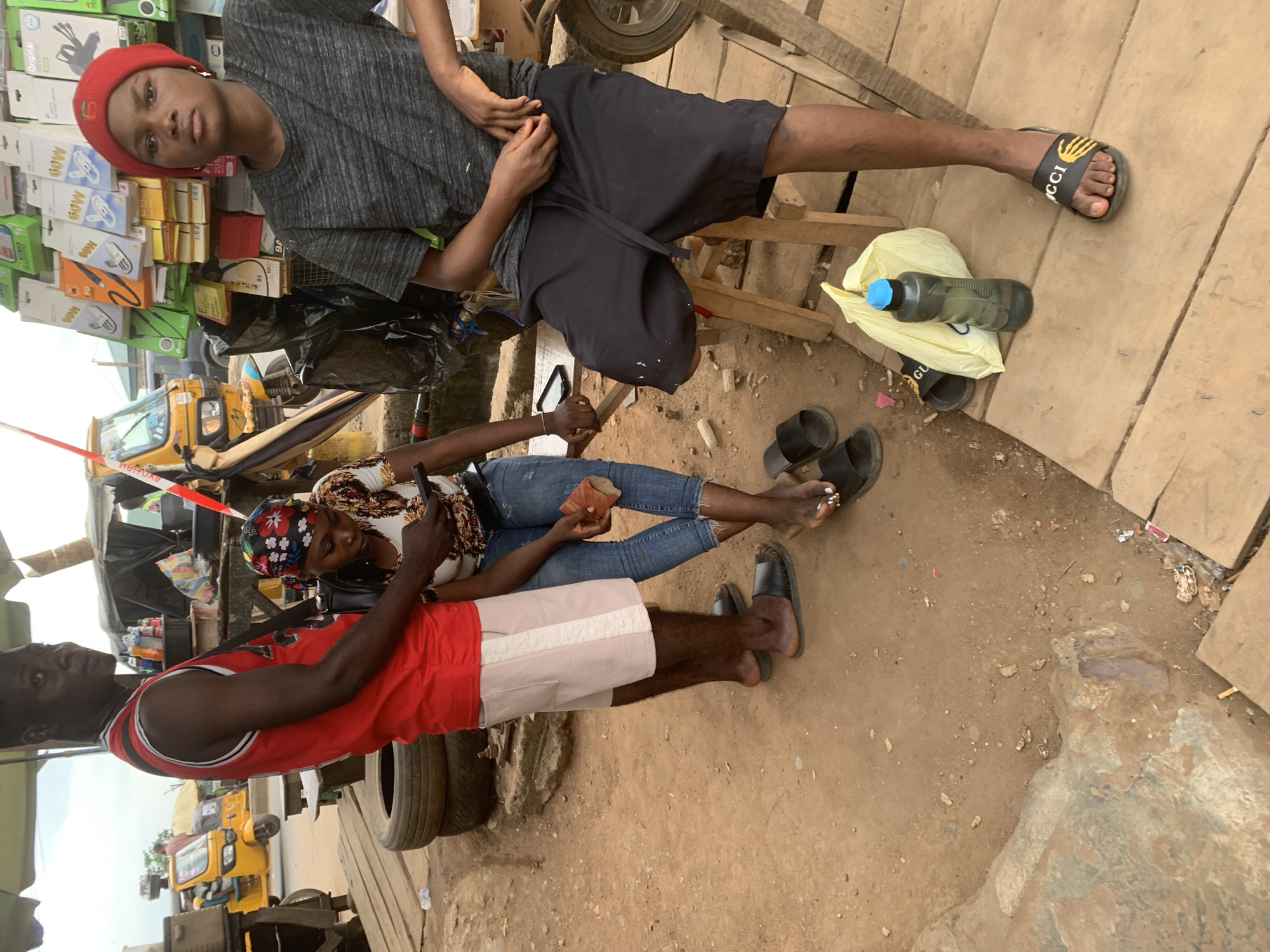A man in a red tank top and white shorts stands to the left of a woman seated on a wooden structure. The man is looking towards the camera, while the woman, wearing a colorful headscarf and a patterned shirt, is looking down at her phone. She is holding a small brown object in her right hand, possibly money. To the right of the seated woman, another person, wearing a grey t-shirt and black shorts, is also seated on a wooden stool. This person is wearing a red beanie and looking towards the camera.
The scene appears to be an outdoor market, likely in Shagamu, Nigeria, given the presence of a yellow motorized rickshaw, commonly known as a Keke Napep, in the background. The ground is dusty and uneven, with several discarded tires stacked to the left of the man. In the background, a stall displays various electronic accessories such as phone chargers and cables. A plastic water bottle with a blue cap and a yellow plastic bag are on the wooden surface in the foreground. The overall atmosphere suggests a bustling commercial area.