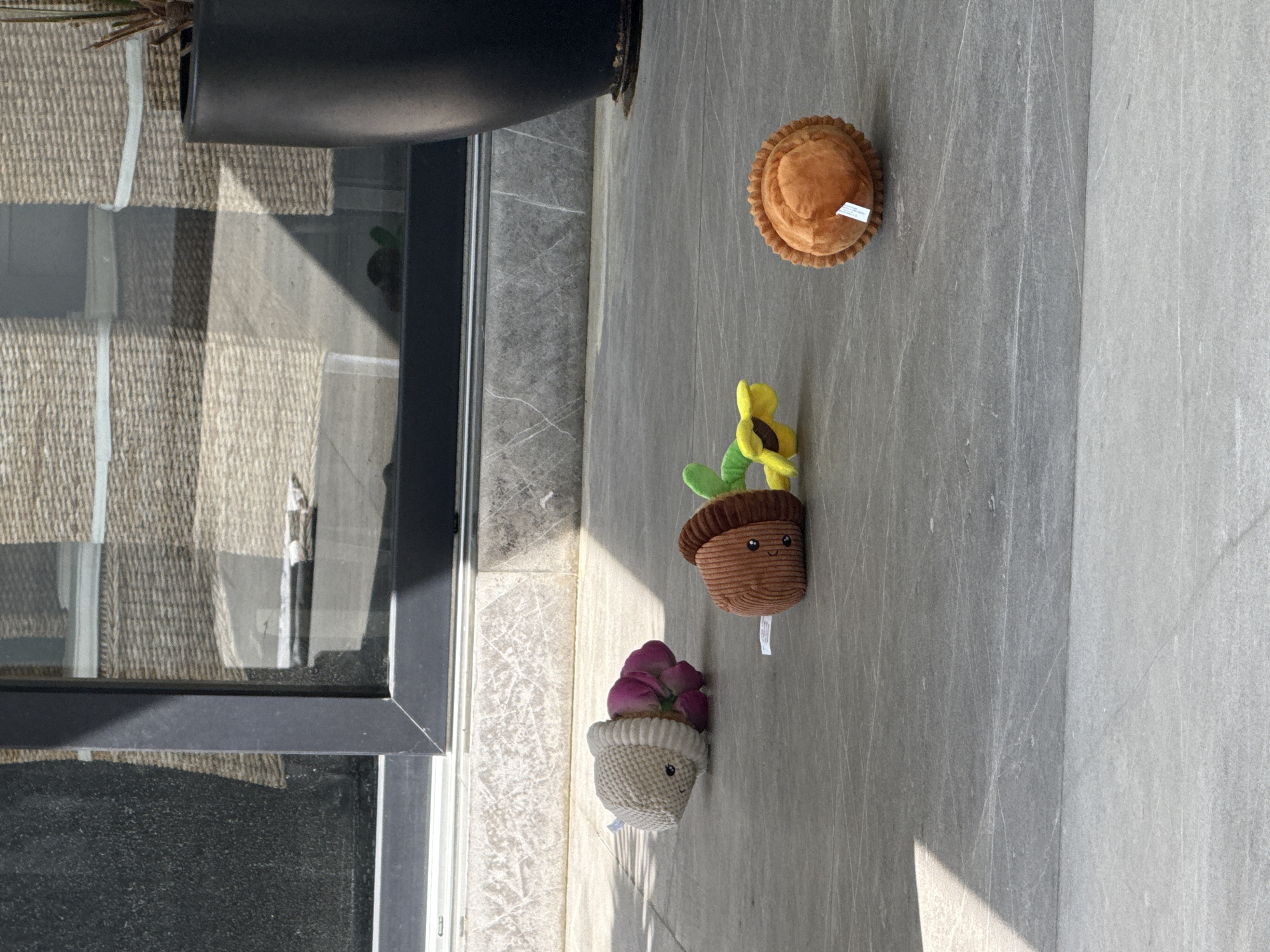 A high-angle shot depicts an indoor setting with a portion of a window or glass door frame visible on the left. A large dark planter sits at the top of the frame. On a gray-toned surface that could be a tile or stone floor, there are four plush toys: a round, pie-like plush toy; a plush toy shaped like a brown pot with a green plant and a yellow sunflower emerging from it; and a plush toy that looks like a pink flower in a white pot. The location is San Bernardino Tlaxcalancingo, Mexico.