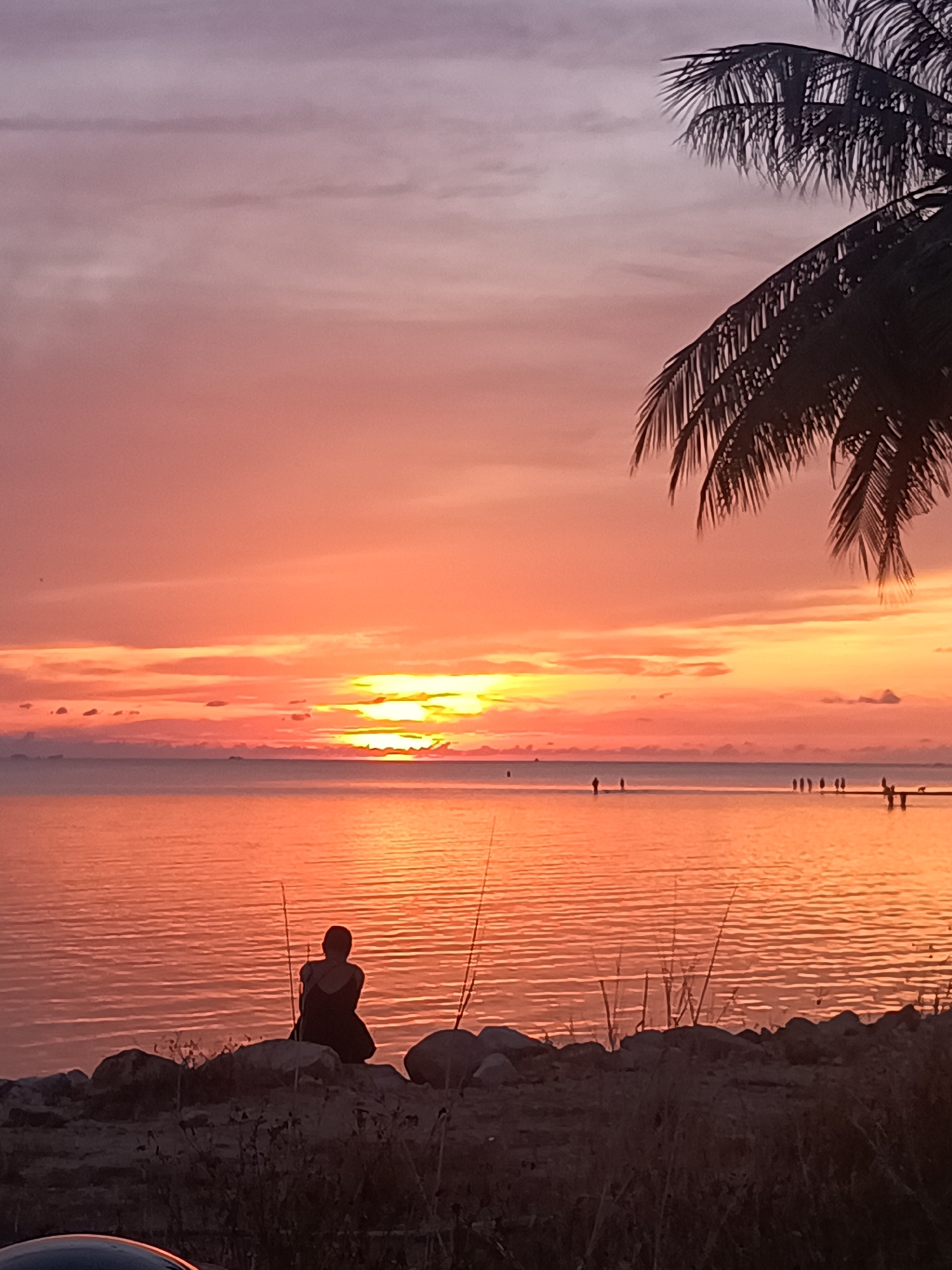 The image shows a sunset scene at a beach in Baan Sri Thanu, Thailand. The sky is a gradient of orange, pink, and purple. The sea reflects the sunset colors. A silhouetted figure of a person is seated on rocks near the shore. Tall grasses and what appears to be fishing poles are also near the rocks. In the distance, silhouettes of people can be seen standing on what appears to be a pier. A palm tree branch is in the top right corner. The bottom portion of what appears to be a vehicle is visible in the bottom left of the scene.
