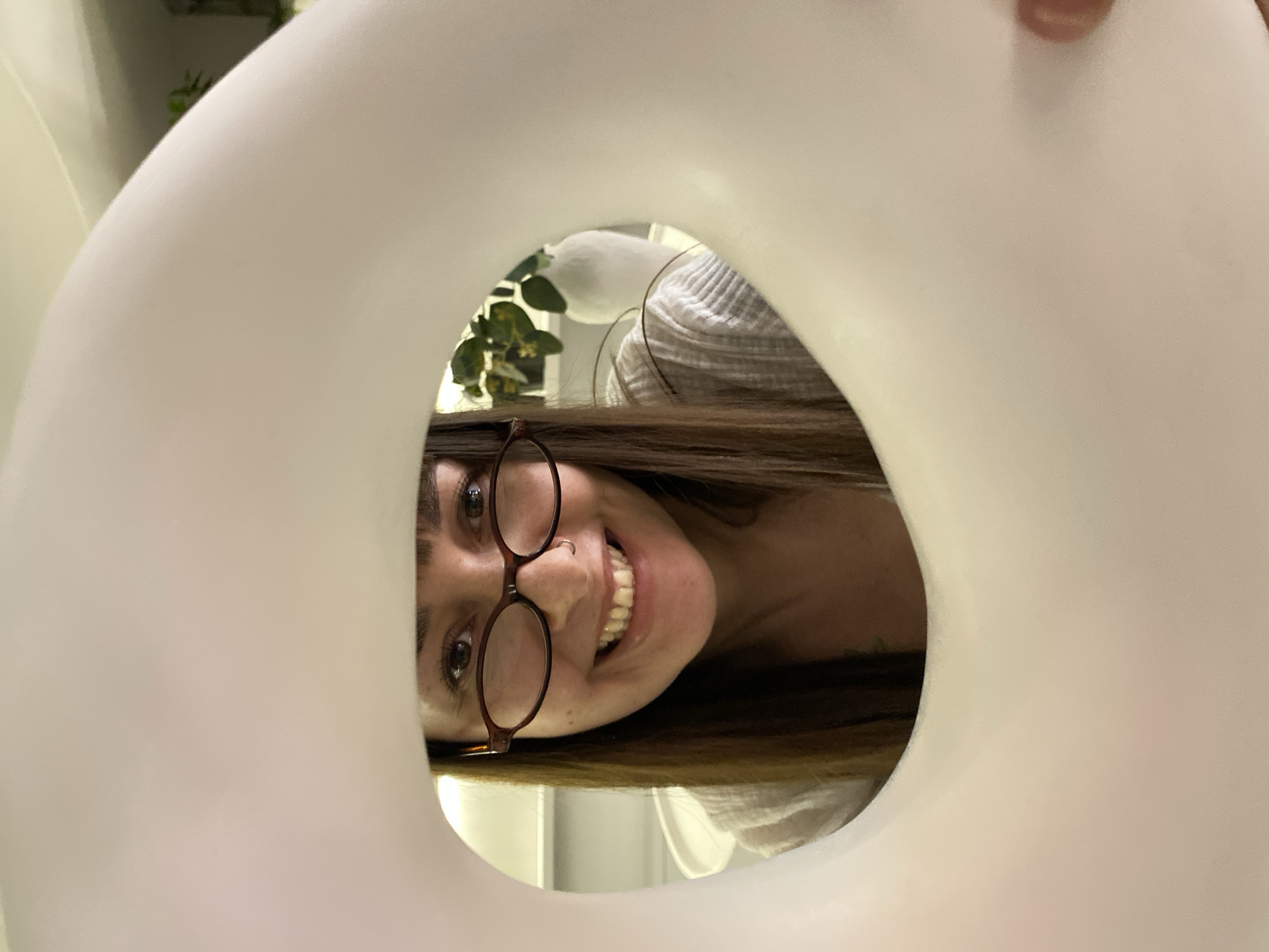 A close-up, eye-level photograph captures a smiling woman peering directly at the camera through the circular opening of a large, soft-looking, white object, possibly an inflatable ring or cushion. The woman has long, dark brown hair and is wearing round, dark-rimmed glasses. A small nose piercing is visible on her right nostril. She displays a wide, cheerful smile, showing her teeth, and her eyes are bright. Her face is positioned horizontally within the frame, suggesting the photograph was taken at a rotated angle. She appears to be wearing a light-colored, possibly striped, garment, visible on her shoulders and upper chest. In the soft-focus background, green foliage from a plant can be seen, indicating an indoor setting. The prominent white object frames the woman's face, creating a playful and intimate perspective. No text is visible in the image.