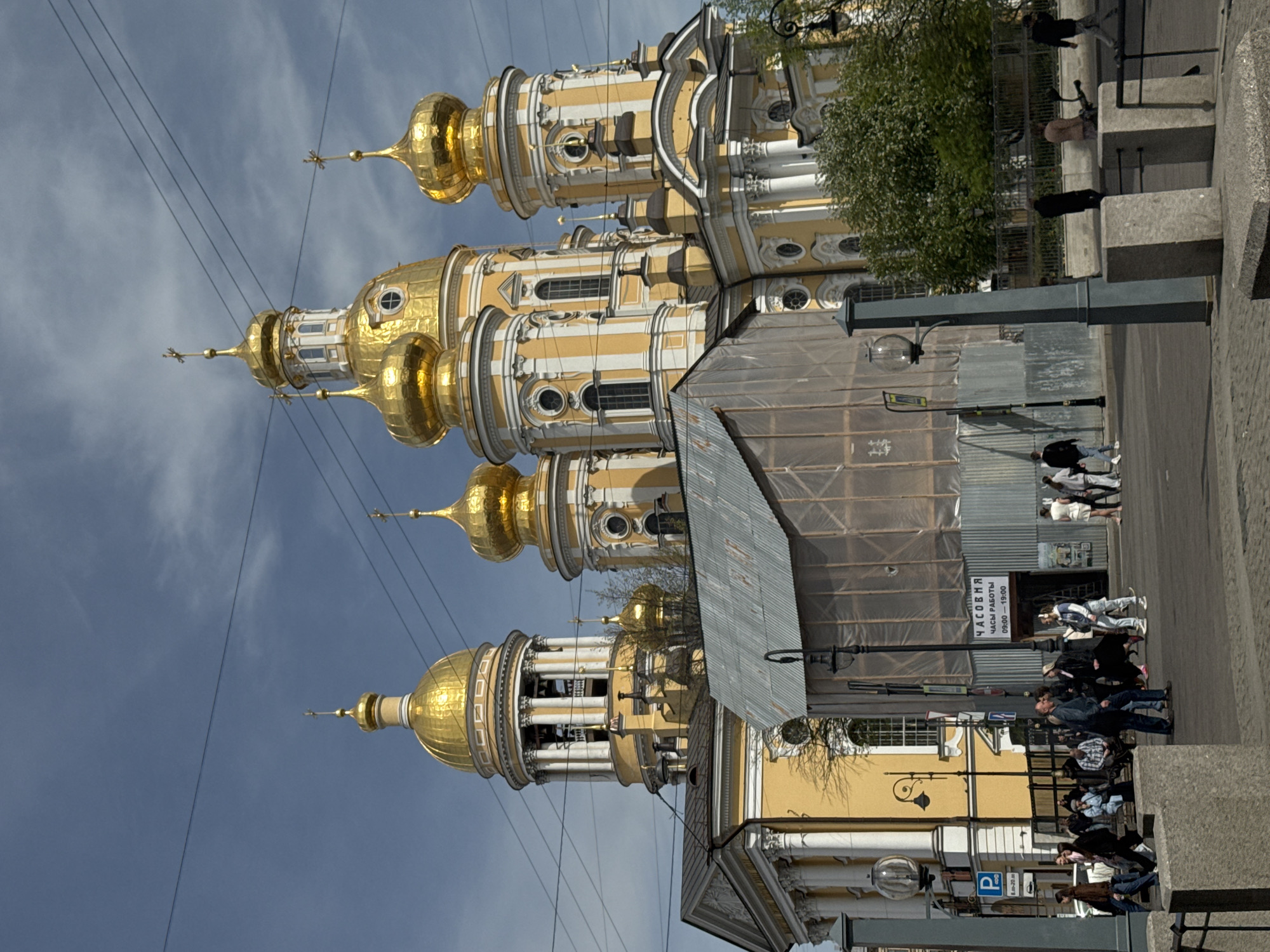 The image depicts the St. Nicholas Naval Cathedral in Saint Petersburg, Russia. The cathedral features ornate gold domes and light yellow facades. In front of the cathedral, a number of people are walking along a sidewalk. The environment suggests a mix of architectural beauty and urban activity. Construction work, possibly a renovation or restoration project, is evident through visible scaffolding.