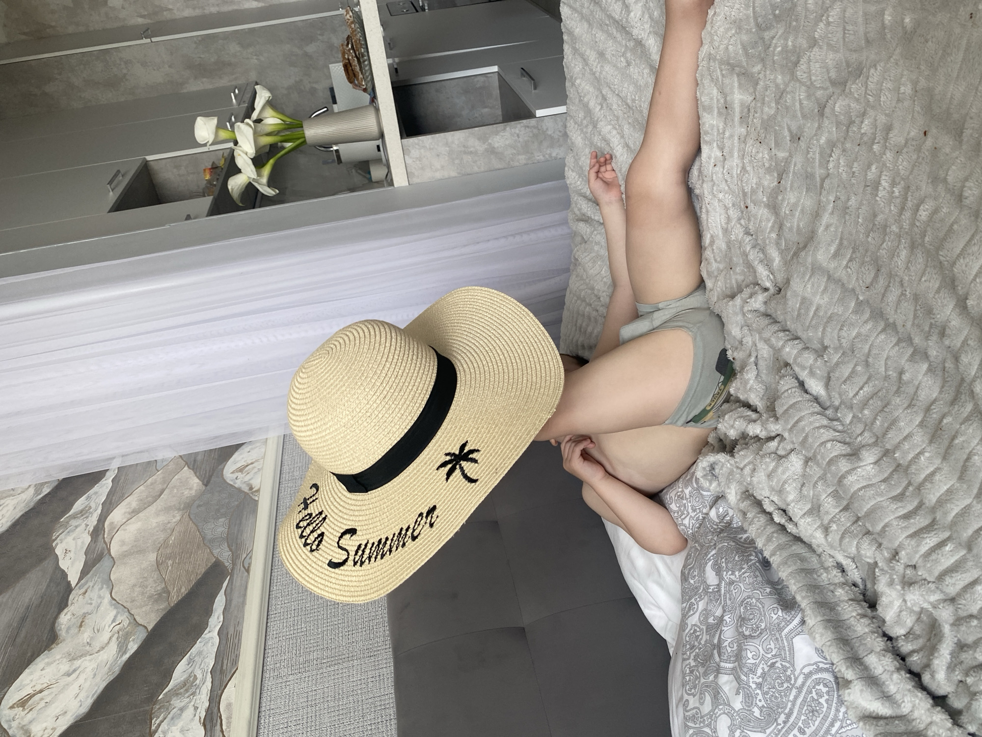 A person is lying on a bed with a large straw hat covering their face. The hat is decorated with the words "Hello Summer" and a palm tree. The person is wearing grey shorts. The bed is covered with a textured grey blanket and a patterned duvet. In the background, there is a grey headboard, a wall with a mountain-themed wallpaper, and sheer white curtains. To the right, a kitchen area is visible with cabinets, a vase of white calla lilies, and a plate of food on a counter. The scene appears to be indoors.