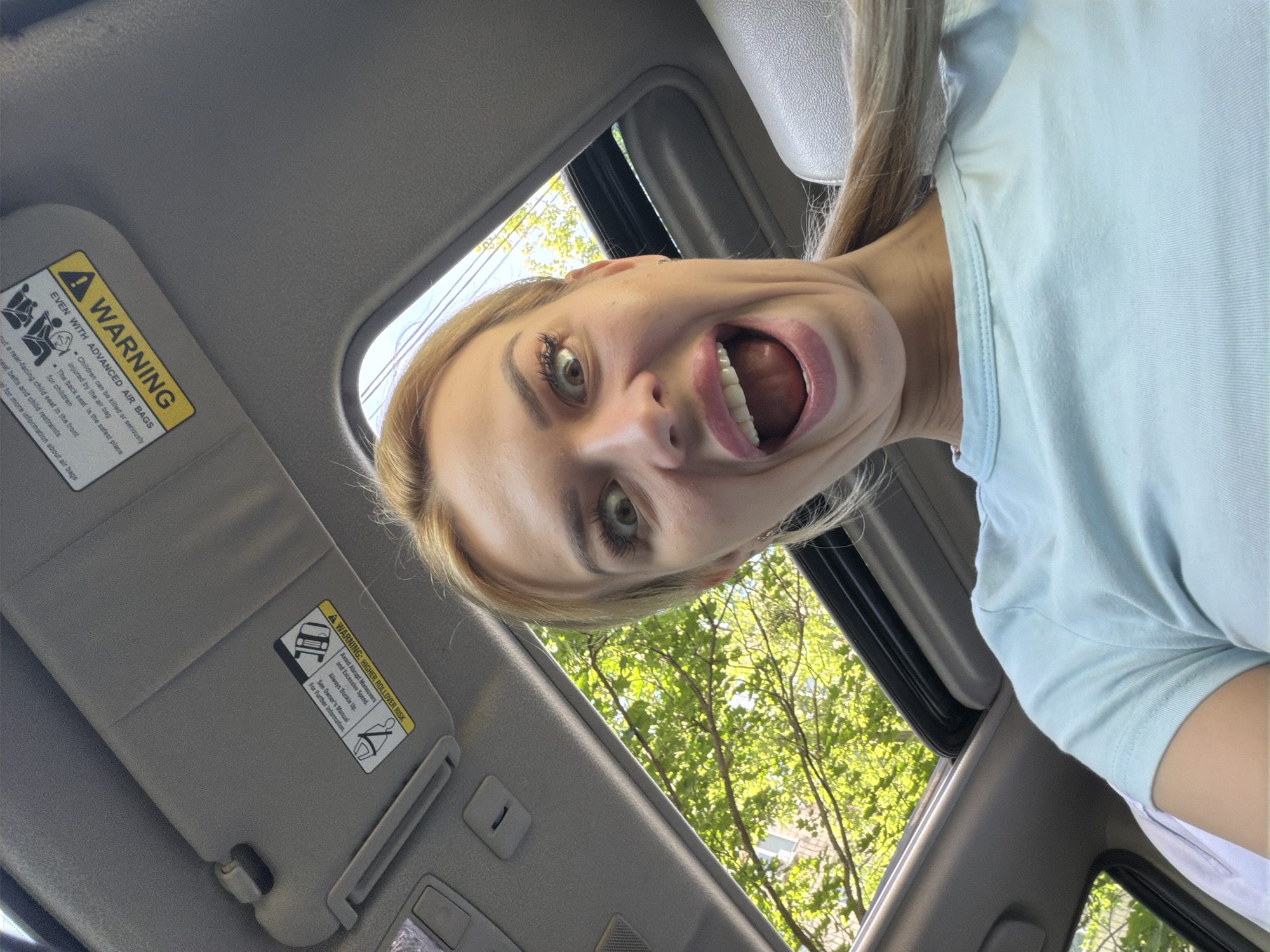 A woman with blonde hair and a light blue t-shirt is looking up at the camera through an open sunroof of a vehicle. She has a wide, surprised expression with her mouth open and eyes wide. The interior of the car is visible, including the gray headliner and a sun visor with warning labels. Outside the sunroof, green trees and a building can be seen. The image appears to be a selfie taken from inside the car. There is no information in the image that places the location in Kyiv, Ukraine.
