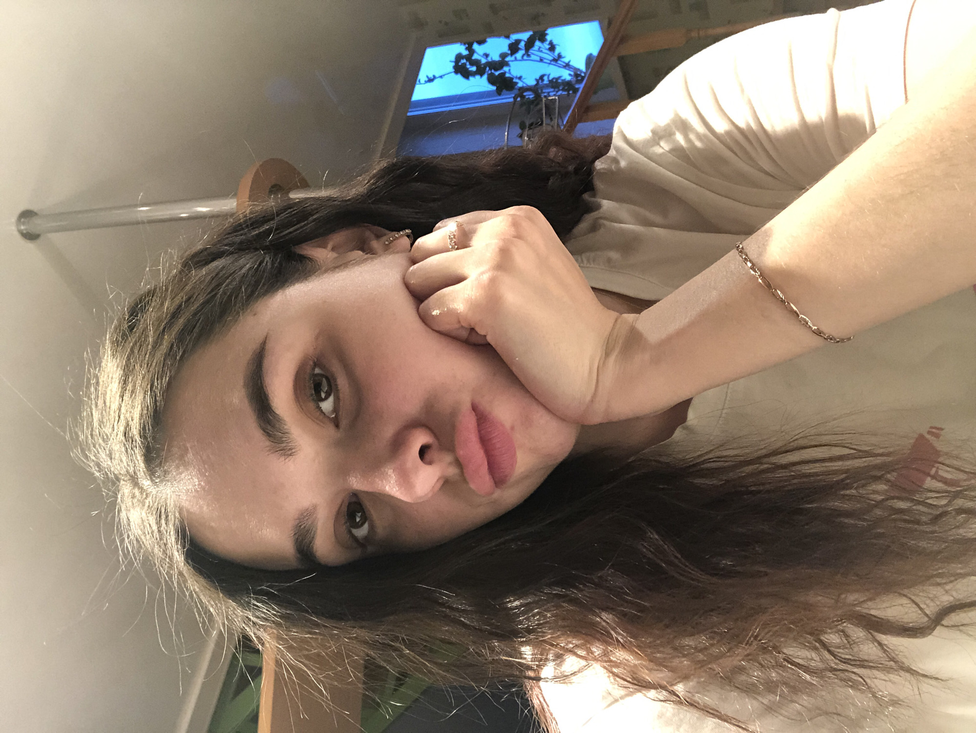 A woman with brown hair is posing for a selfie indoors. She is lying down with her head resting on her right hand, wearing a light-colored shirt and a delicate gold bracelet on her left wrist. A ring is visible on her right hand. In the background, there is a window showing a tree and a blue sky. There is also a horizontal pipe with a wooden attachment visible near the top left corner. The image is rotated slightly counterclockwise. This image cannot be located in any specific location including the city of Noviy, Russia.