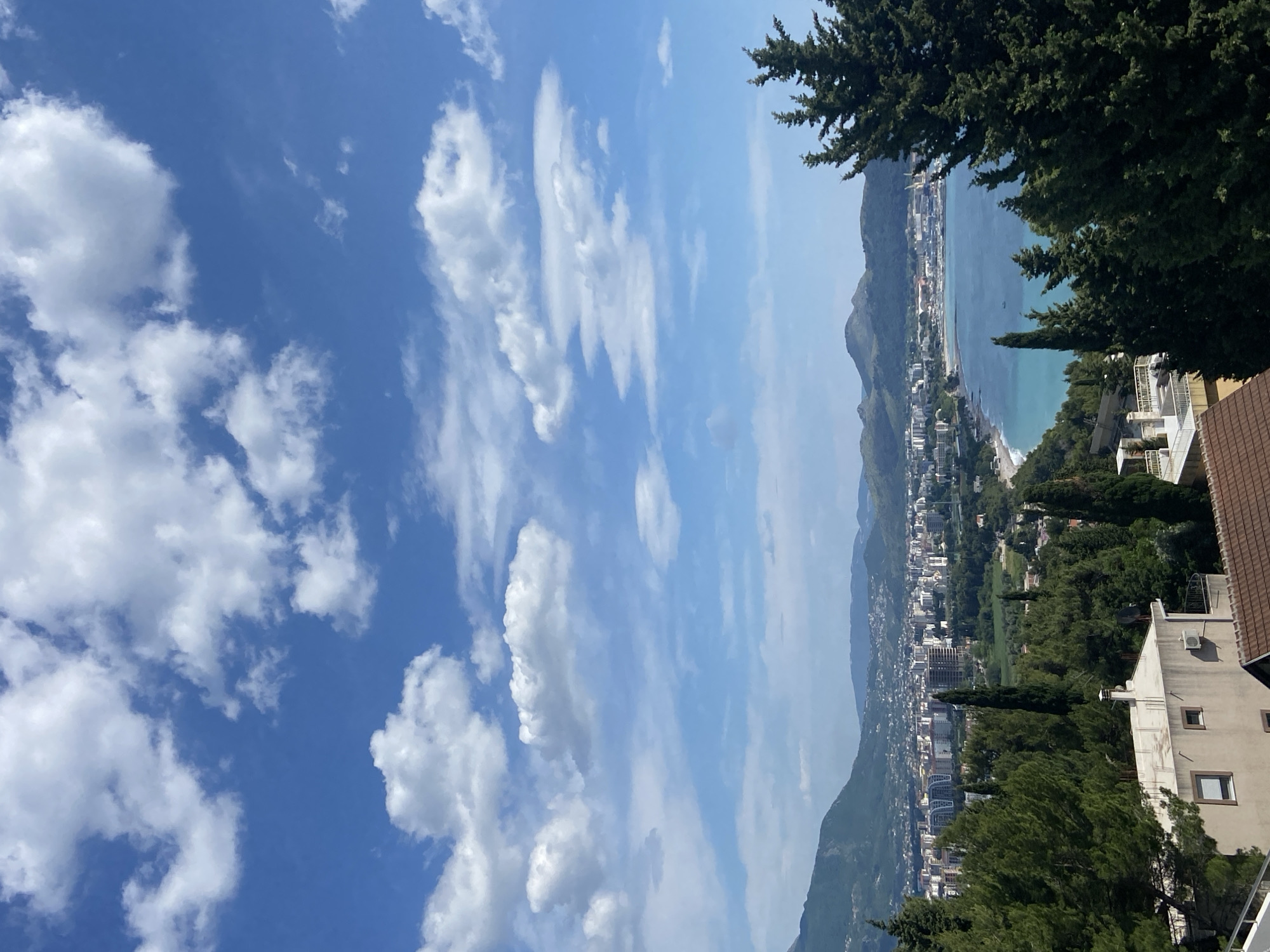 The image depicts a coastal cityscape, likely Šušanj, Montenegro, viewed from an elevated perspective. The scene is dominated by a bright blue sky with scattered cumulus clouds. In the middle ground, a coastline with a sandy beach borders turquoise waters. The city extends along the coast, with buildings of varying heights and architectural styles intermixed with green spaces and trees. Mountains rise in the background, partially obscured by atmospheric haze. In the foreground, parts of buildings with tiled roofs and dense green foliage, including coniferous trees, frame the view. There are no visible people.