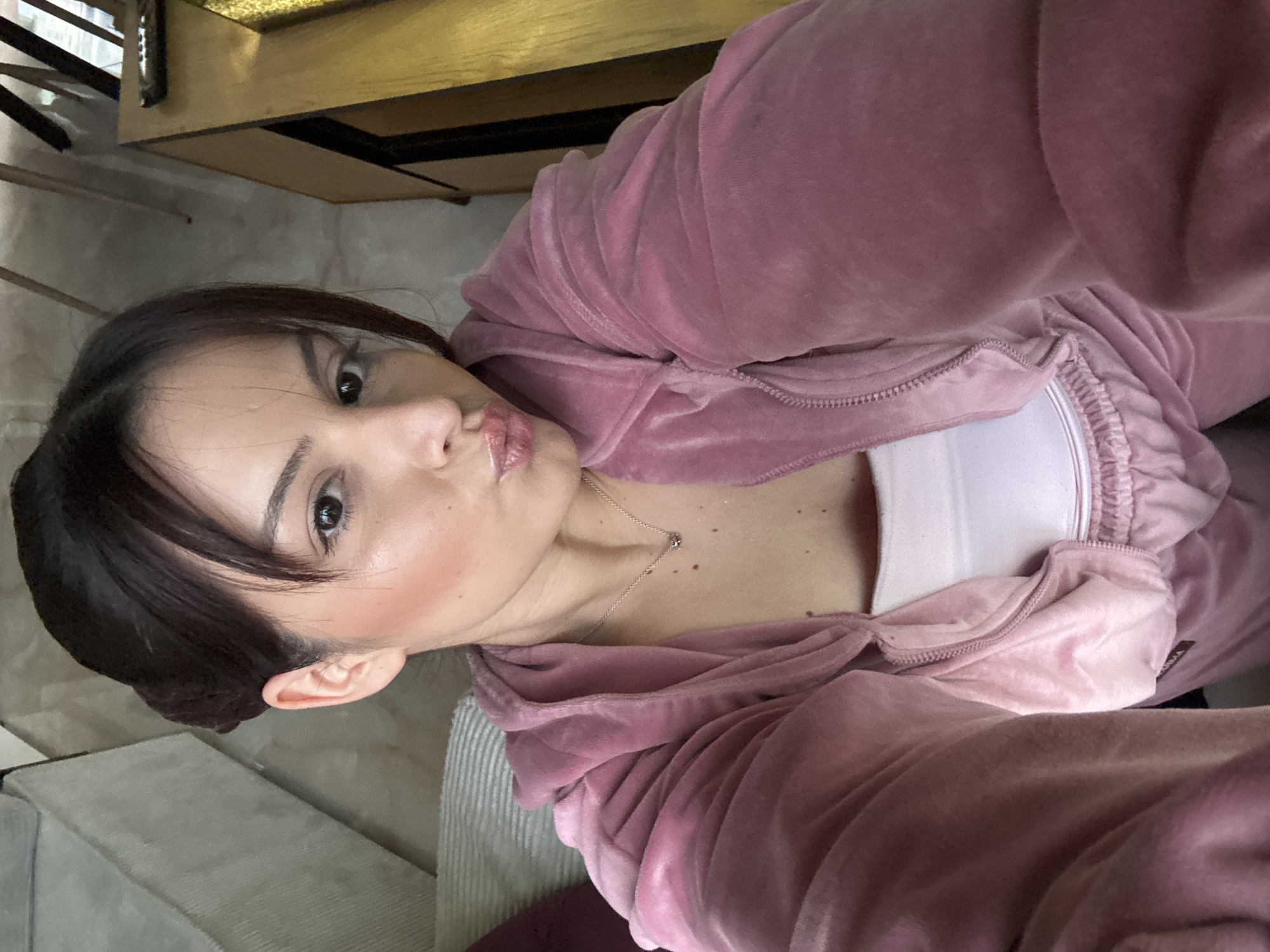 A woman with dark hair pulled back and wearing a pink velour tracksuit is posing for a selfie. She is looking at the camera with a slight pout. She is wearing a white top underneath her tracksuit. A silver necklace is visible on her neck. The background is blurry and includes a wooden piece of furniture and part of a light-colored sofa. There is no indication of Burgas, Bulgaria in the image.