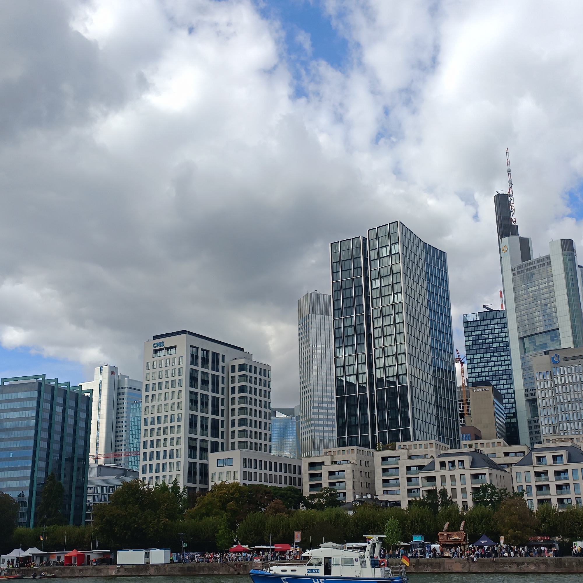 The image captures a cloudy day in Frankfurt, Germany, showcasing the city's modern skyline. The foreground features the Main River, with a police boat docked near the bank. Several buildings of varying architectural styles line the river, including the prominent CMS Tower, identifiable by its signage. Dense trees separate the riverfront from the cityscape, with a crowd of people gathered along the embankment, suggesting a public event or gathering. Tents and food stalls are visible, further indicating a lively atmosphere. The background is dominated by a collection of high-rise buildings, characteristic of Frankfurt's financial district, with a mix of glass and concrete facades. The sky is overcast with dramatic cloud formations, casting a diffused light over the scene.
