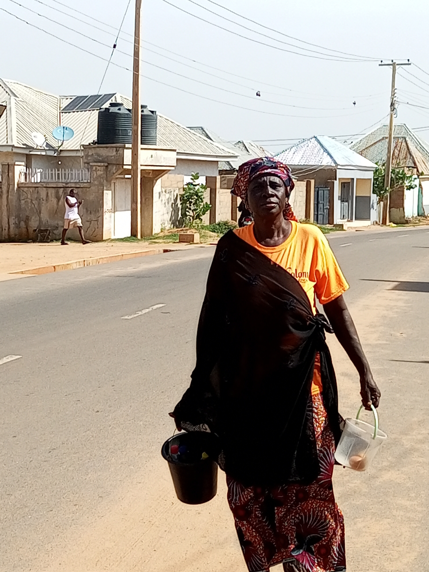 A woman walks down a paved street in Tempure, Nigeria, carrying two buckets. She wears a red, patterned headscarf, an orange t-shirt with the word "Colonial" visible on it, and a patterned skirt. A dark, flowing fabric is draped over her shoulders. In her right hand, she carries a clear plastic bucket containing at least two brown eggs. In her left hand, she holds a black bucket filled with various colorful objects, possibly items for sale. The street is lined with modest houses with corrugated metal roofs. In the background, a man in a white shirt and shorts stands on a sidewalk, looking at his phone. Several power lines are visible overhead, supported by wooden poles. The sky is clear and bright.