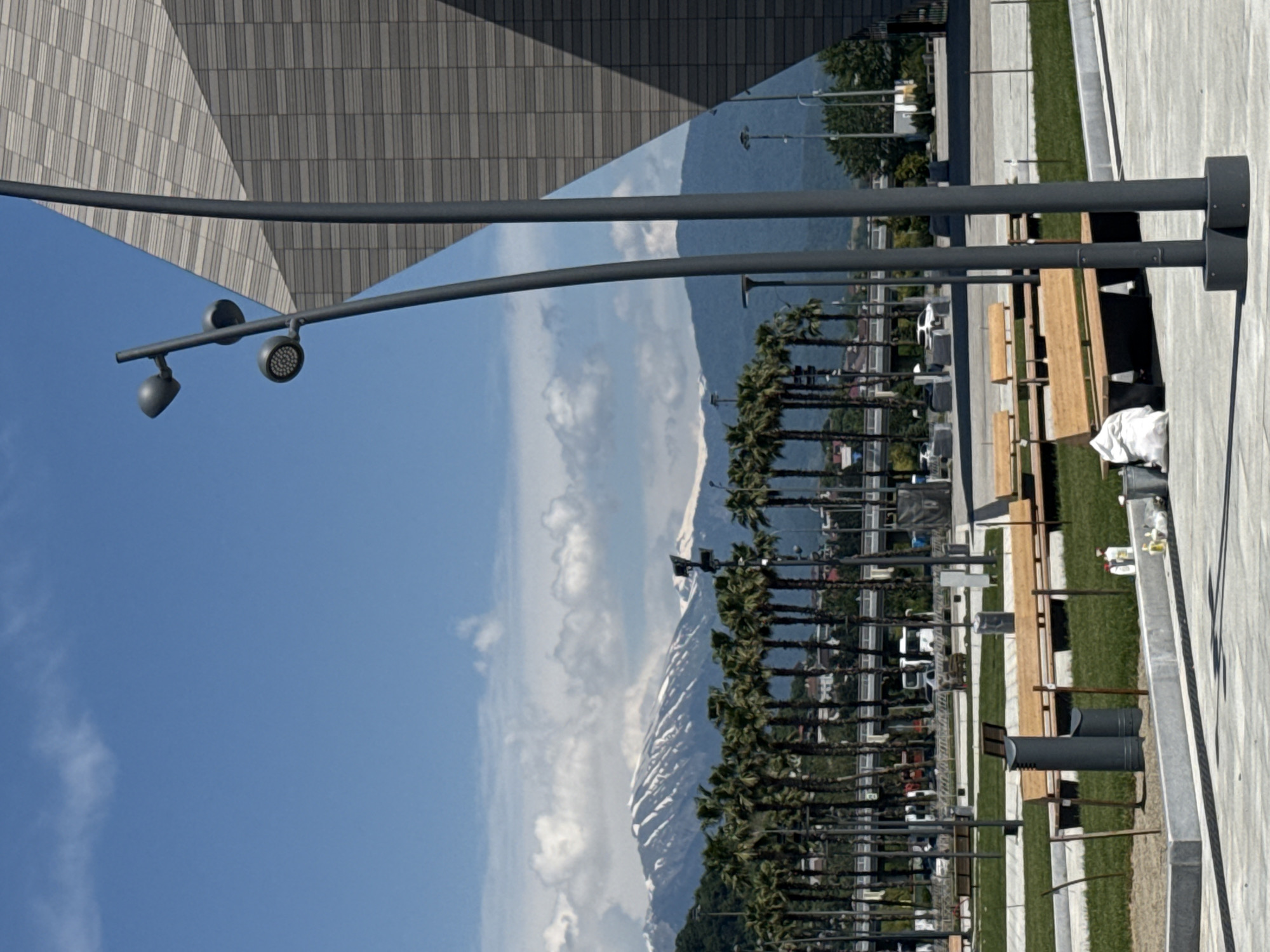 The image depicts an outdoor scene in Sirius, Russia, likely within the Olympic Park area, characterized by modern urban planning and a mountainous backdrop. In the foreground, two dark grey, cylindrical street lamp poles with octagonal bases are prominently positioned on a light grey paved surface. The left pole features three curved arms, each terminating in a circular LED light fixture. To the left of these poles, a grey cylindrical trash receptacle and a smaller bin are visible next to a light wooden bench with a dark base, situated on a grassy area. Nearby, a grey bucket and several plastic bottles (two white, one clear) are on the pavement. Further right, a white bag rests on the pavement near another wooden bench.
The mid-ground consists of a landscaped park area with meticulously maintained green grass and multiple light wooden benches with dark supports. Behind this park space, a long, dense row of tall palm trees with green fronds extends horizontally across the frame. Beyond the palm trees, an elevated concrete road or bridge structure is visible, populated by several vehicles including white cars, vans, and a bus. Various standard streetlights are interspersed along this elevated structure and among the palm trees.
The background is dominated by a majestic range of snow-capped mountains, displaying distinct patterns of white snow against darker rock. The sky above is a clear, vibrant blue with scattered white cumulus clouds. To the far right, a large, modern building with a geometrically textured facade is partially visible. The building's surface consists of rectangular panels arranged in alternating patterns of light and dark grey, creating a striking architectural element. The scene is illuminated by bright daylight, casting clear shadows and indicating sunny weather. No people are clearly discernible in the image.