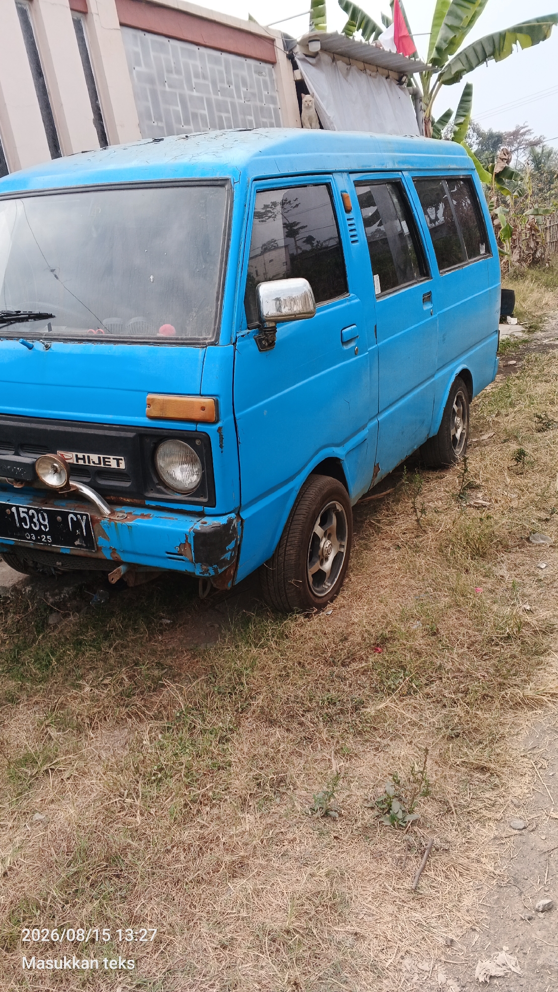 A blue Daihatsu Hijet van is parked on a dry, grassy area. The van has a license plate that reads "1539 CY" and "03-25". There is some rust visible on the front bumper and body of the van. In the background, there is a building with a striped facade and some vegetation, including banana trees. A red and white flag is visible on top of the building. The sky is overcast. The timestamp "2026/08/15 13:27" and text "Masukkan teks" are overlaid on the lower portion of the image.