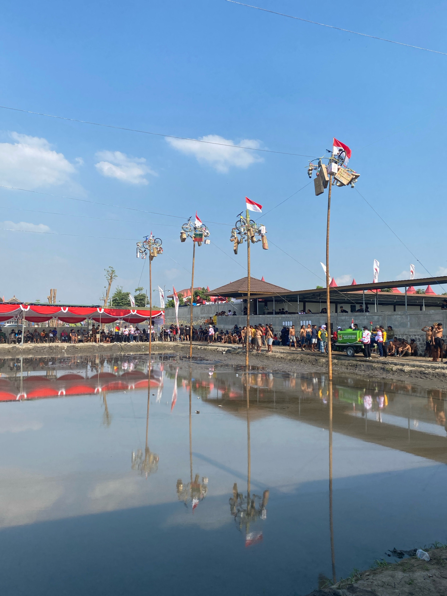 This image depicts a scene of a festive gathering, possibly a competition or celebration, taking place outdoors in Kediri City, Indonesia. The foreground is dominated by a large body of water, likely a pond or shallow lake, with calm, reflective surfaces mirroring the sky and the structures on the far bank.
Along the edge of the water, a crowd of people is gathered. They are positioned behind a line of tents with red and white awnings, suggesting a designated viewing area. Many of the onlookers are seated or standing, observing the events unfolding.
In the middle ground, several tall poles are erected. Each pole is adorned with decorations, including what appear to be Indonesian flags (red and white), bicycles, and various other objects. These poles serve as central focal points of the scene, hinting at a competition or display.
Further back, a building with a prominent brown roof is visible, providing shade and a backdrop to the activity. More tents and structures line the area behind the main gathering. A small green vehicle, possibly a utility cart or stage prop, is also present in this area, with people gathered around it.
The sky is a clear blue with scattered white clouds, indicating fair weather. The overall impression is one of a lively community event, likely celebrating a holiday or tradition, with a strong sense of local participation and pride, as evidenced by the Indonesian flags. The reflection of the poles and flags in the water adds a unique visual element to the composition.