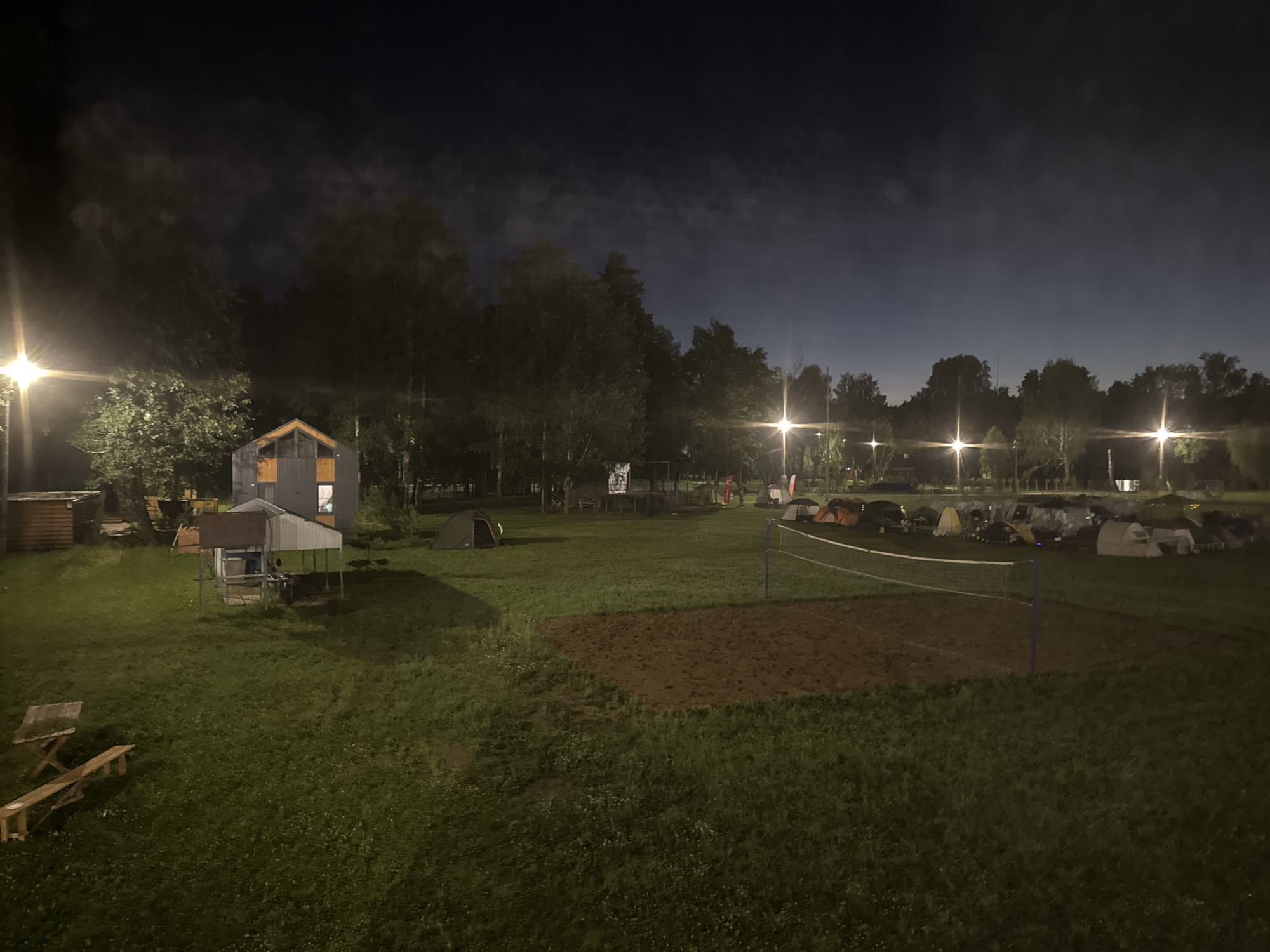 The image depicts a night scene at an outdoor campsite, potentially located in the vicinity of Doma 1096 Km, Russia. A row of tents is visible in the background, set up on a grassy field. A volleyball net and a sand court suggest recreational activities. A small, open-sided structure, possibly a shelter or picnic area, is situated in the middle ground, with a picnic table and bench nearby. In the far left, a wooden structure resembling a small building or shed is present. Several artificial lights illuminate the scene, including streetlights and lights from within buildings. The sky is dark, with some faint clouds. No people are clearly visible in the image.