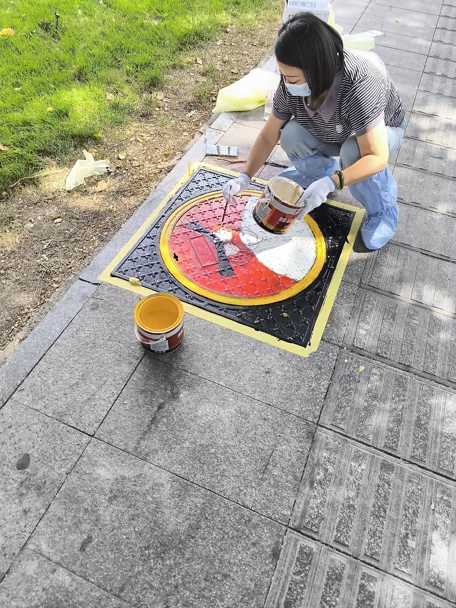 In Ürümqi, China, a woman wearing a face mask, gloves, and protective shoe covers is crouched on a sidewalk, painting a circular design on a manhole cover. She is holding a can of paint and a brush, and appears to be applying white paint to a red and white pattern on the cover. Two open paint cans are visible on the sidewalk nearby, one yellow and one with a red and white label that reads "精选" (jingxuan, meaning "selected" or "premium"). The manhole cover is surrounded by yellow tape, and the surrounding sidewalk is made of grey paving stones. To the left of the sidewalk, a patch of green grass with some fallen leaves is visible.